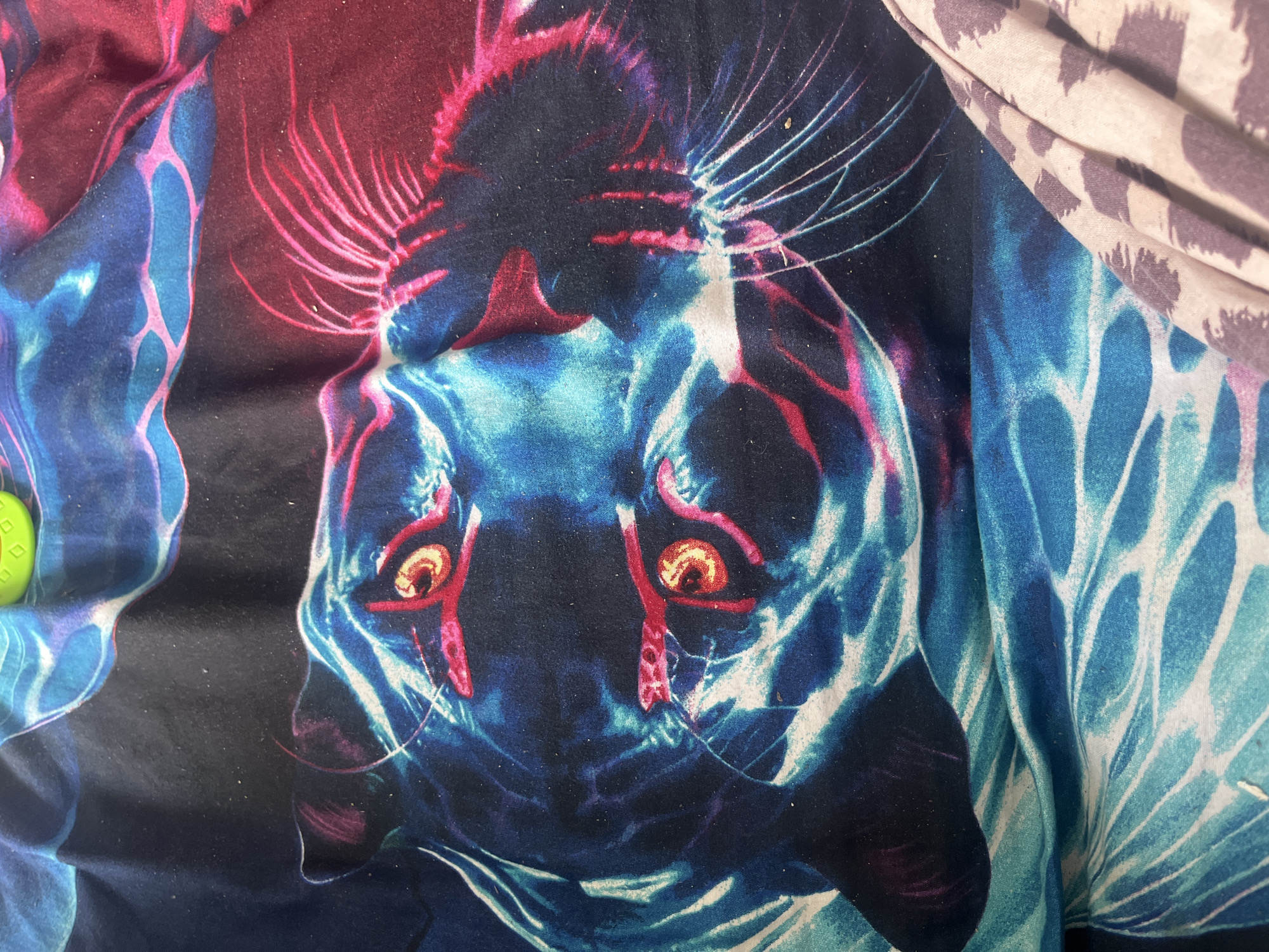 The image is a close-up of a fabric with a vibrant, stylized print of a feline face, likely a tiger or panther, rendered in electric blue, neon pink, and deep crimson. The eyes are a prominent feature, depicted as glowing orange with red pupils, surrounded by bright pink outlines. The fur is suggested with swirling patterns of blue and white, and the whiskers are sharp, fine lines of pink and white radiating outwards. The fabric appears to be creased and folded, creating shadows and highlighting the texture. A small, green, diamond-patterned object is partially visible in the upper left corner, its surface catching the light. In the lower right, a portion of another fabric with a muted, spotted pattern is visible, creating a contrast with the main subject. There is no indication of the location in the city of Chelyabinsk, Russia, nor any people, actions, or interactions. The scene is a static depiction of a printed textile.