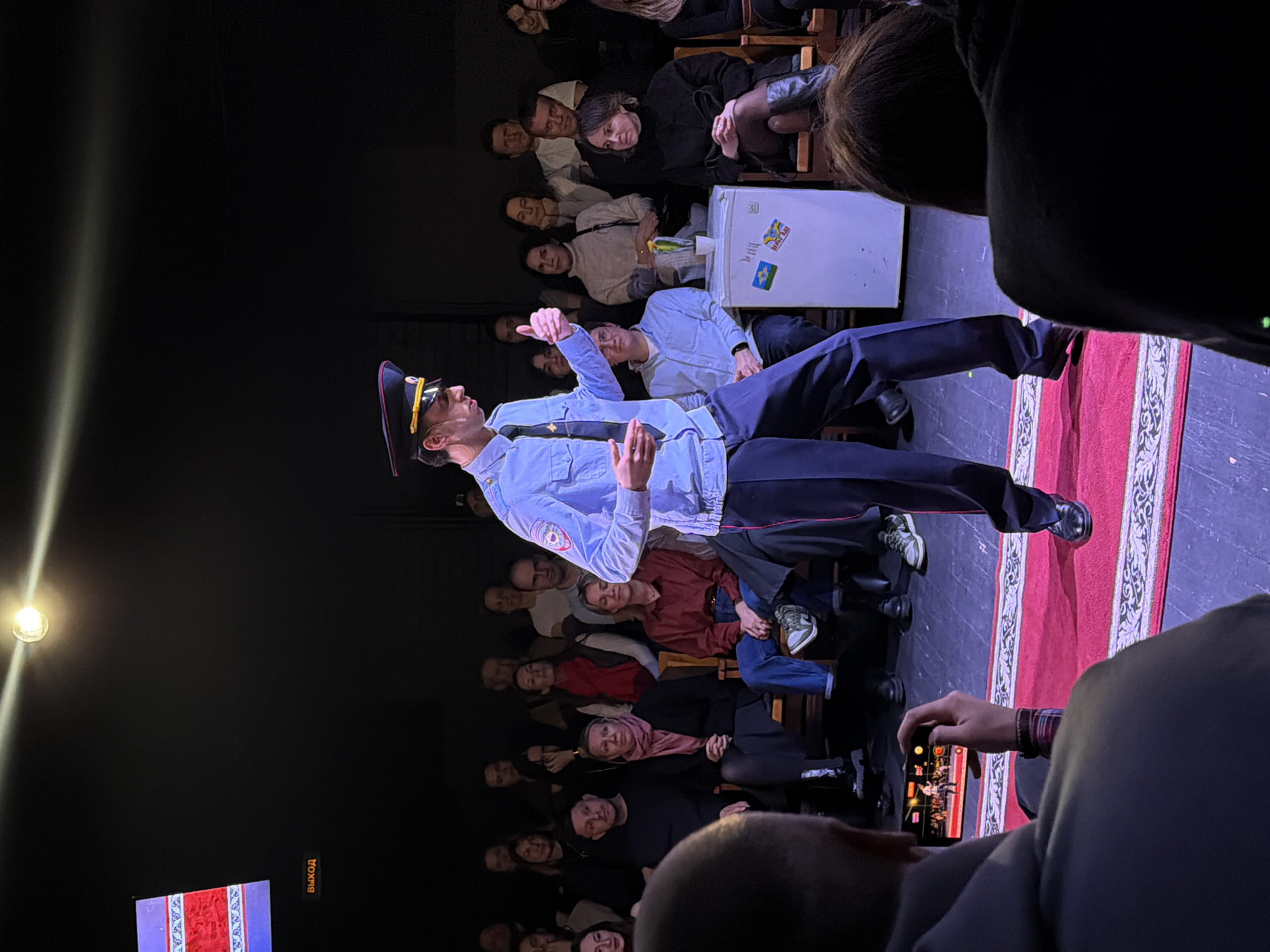 This media file captures an indoor performance or event, viewed from the audience's perspective, likely in Moscow, Russia, given the provided context and visible Cyrillic text.
The main subject is a male performer on stage, dressed in what appears to be a light blue uniform shirt with epaulets, a dark tie, dark trousers, and a peaked cap with a badge, possibly representing a police or military figure. He wears sunglasses and is standing confidently, gesturing with a thumbs-up with his right hand, looking towards his left (the audience's right).
On the stage, behind the main performer, is a small, white, top-opening refrigerator, serving as a prop. Another man in a light blue shirt and dark trousers is seated on this refrigerator, looking towards the main performer. The refrigerator has several stickers, including one that strongly resembles the Ukrainian flag (blue and yellow stripes) and another with the Cyrillic word "ВЫХОД" (Exit) visible, rotated. A patterned red carpet or runner is laid out on the stage floor.
A diverse audience is seated in rows, facing the stage. Their faces are visible as they watch the performance, with varying expressions suggesting engagement and observation. In the bottom right foreground, an audience member is holding a smartphone, appearing to record the event on stage.
The venue is dimly lit, with a bright spotlight visible on the far left, illuminating the stage. The overall impression is that of a theatrical or comedic performance. The presence of the "ВЫХОД" text in Cyrillic on the stage prop reinforces the Russian-speaking location.