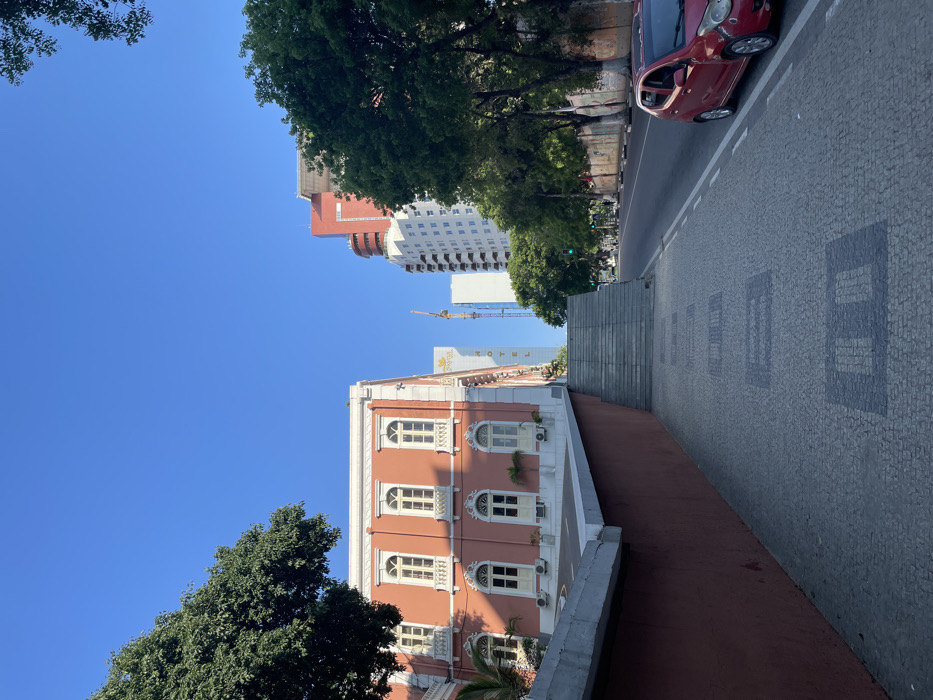 The image, originally rotated 90 degrees counter-clockwise, captures a sunny urban street scene in Luanda, Angola. In the foreground, an elegant, multi-story building with terracotta-colored walls and ornate white architectural trim stands prominently on the left. It features several arched windows, some adorned with small potted plants or air conditioning units.
Adjacent to this building, a wide pedestrian area or sidewalk is paved with intricate gray cobblestones forming distinct square patterns, characteristic of Calçada Portuguesa. A flight of stone steps ascends from this area towards the upper right. Further to the right, a paved road is visible, where a bright red car, likely a compact hatchback such as a Fiat 500, is parked by the curb.
Lush green trees are scattered across the scene, framing the foreground and lining the street in the mid-ground, casting shadows. In the background, modern high-rise buildings rise against the clear blue sky. One tall building features a distinctive red upper section, while another, predominantly white structure, clearly displays the word "HOTEL" on its facade. A yellow construction crane is visible between these taller buildings, suggesting ongoing development in the area.
The bright, direct sunlight and deep blue sky indicate a clear, sunny day, likely mid-morning or early afternoon, creating a vibrant and distinct atmosphere characteristic of a city with Portuguese colonial heritage.