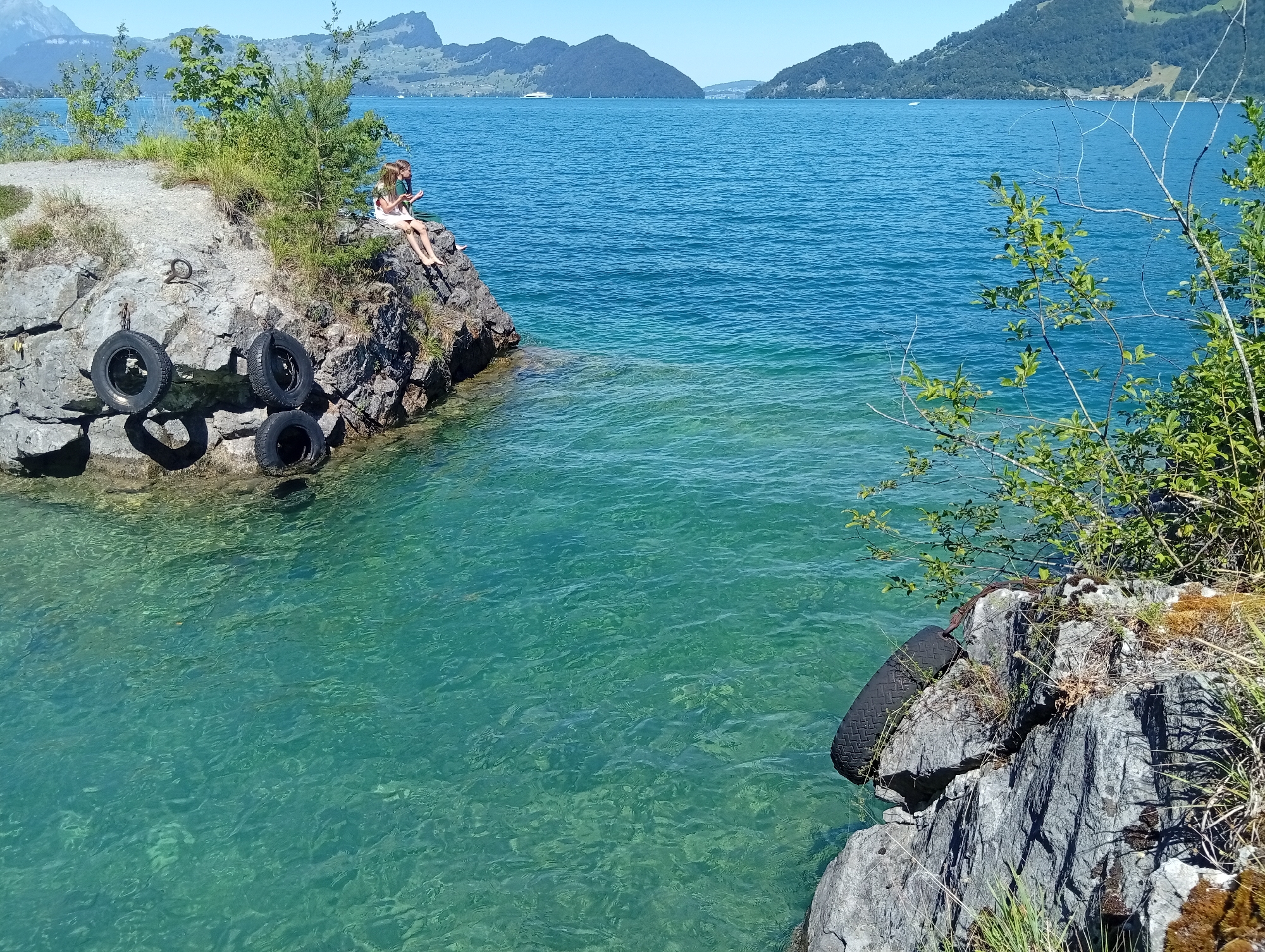 Two young people, a girl and a boy, are sitting on the edge of a rocky outcrop overlooking a clear blue lake in Emmetten, Switzerland. The girl, wearing a white dress, and the boy, in a green shirt, are facing the water with their legs dangling. Three black tires are visible on the rocks near the water's edge. In the background, mountains rise behind the lake, and the sky is clear and blue. The water is calm and transparent, revealing the rocky bottom below. The scene depicts a peaceful summer day at the lake.