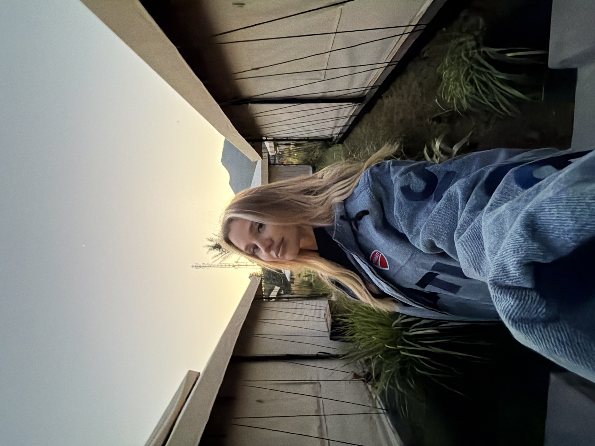 A blonde woman with her hair down is taking a selfie outside in a glamping site. She is wearing a blue denim jacket with a red Ducati logo on the left breast. The woman is standing between two canvas tents. The tents are beige and have black ropes attached to them. There are green plants in front of the tents. The sky is a light blue with hints of orange, suggesting it is either sunrise or sunset. In the background, mountains can be seen. This location is identified as Kintamani, Indonesia.