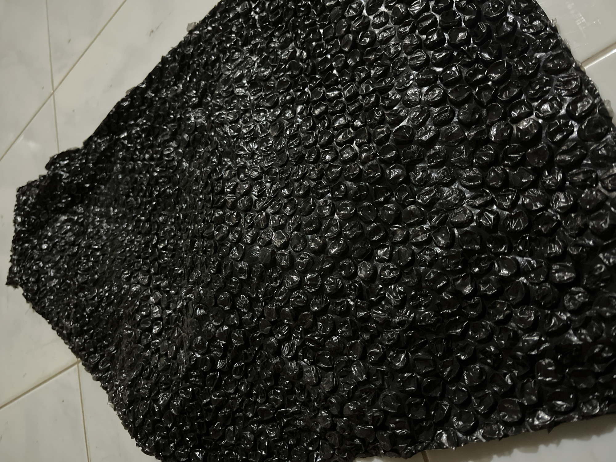 The image shows a sheet of black bubble wrap spread out on a light-colored tiled surface. The bubble wrap fills most of the frame, with its textured surface of raised bubbles reflecting light and creating a shiny, crinkled appearance. The lighting appears to be artificial or overcast, as there are no strong shadows. The tiled surface, visible in the upper left and lower right corners of the image, suggests an indoor setting, possibly a floor. There are no people or animals present, and the focus is solely on the material and its texture. The image is a still shot, with no action or event depicted. The overall impression is a close-up view of the bubble wrap, highlighting its materiality. The location context of Kota Kuala Simpang, Indonesia, is noted but does not provide any visual cues within the image itself.