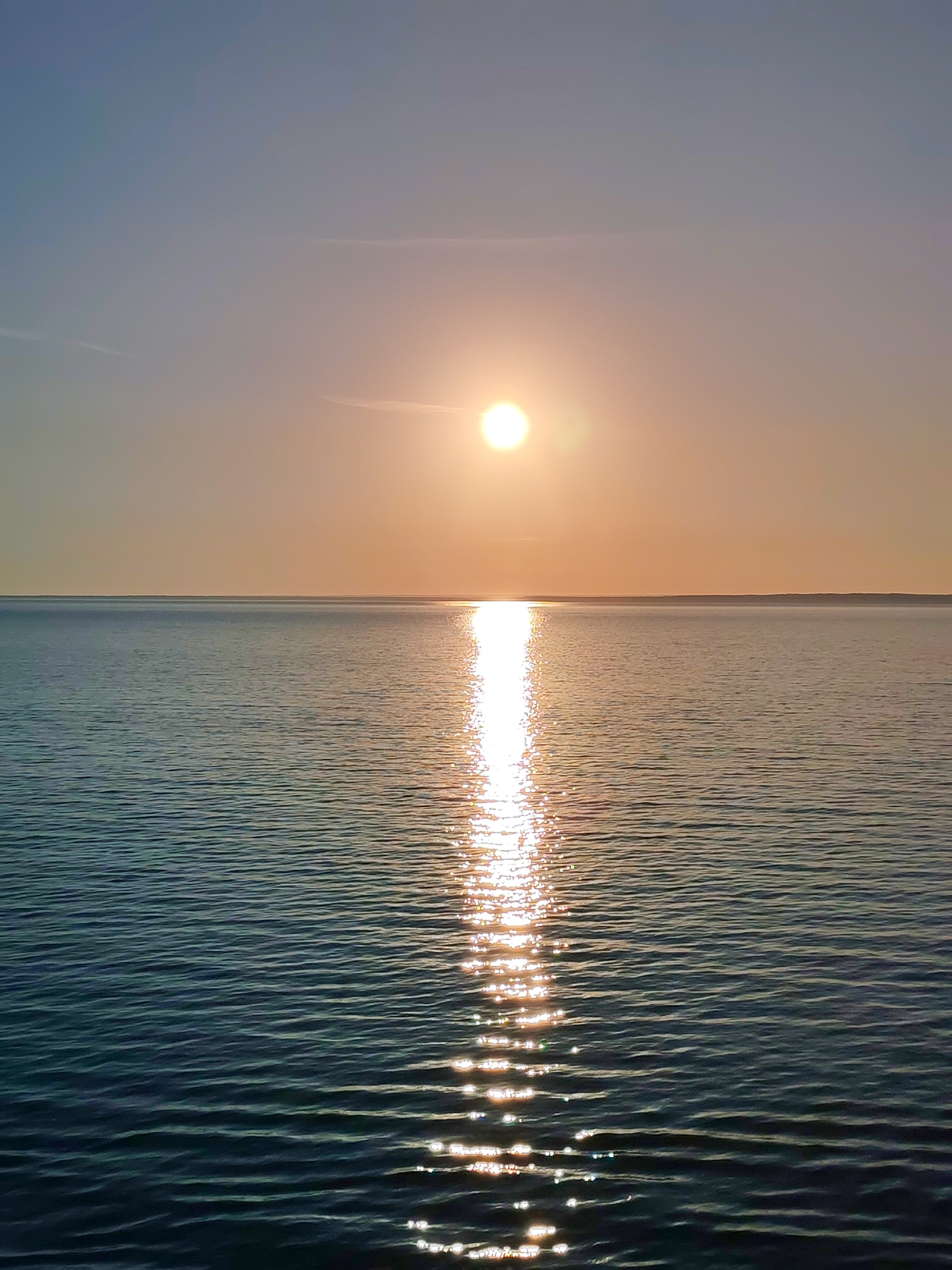 The image depicts a sunset over a body of water, likely a lake or sea, in Ozernoe Ust'e, Russia. The sun is positioned near the center of the frame, casting a bright, vertical reflection on the water's surface. The sky is a gradient of light blues and oranges, transitioning to a brighter yellow-orange near the sun. The water's surface shows small waves that create a textured reflection of the sunlight. In the distance, a dark, flat land mass can be seen.