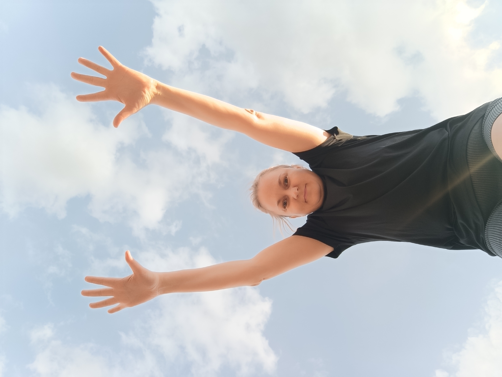 A low-angle shot depicts a woman from a worm's eye view against a backdrop of a partly cloudy, bright sky. The woman has fair skin and blonde hair pulled back. She wears a black t-shirt and dark shorts. Her arms are extended outwards and upwards, with her hands open and facing the camera. The location is not discernible as Dubovaya, Russia, solely from the visual content.