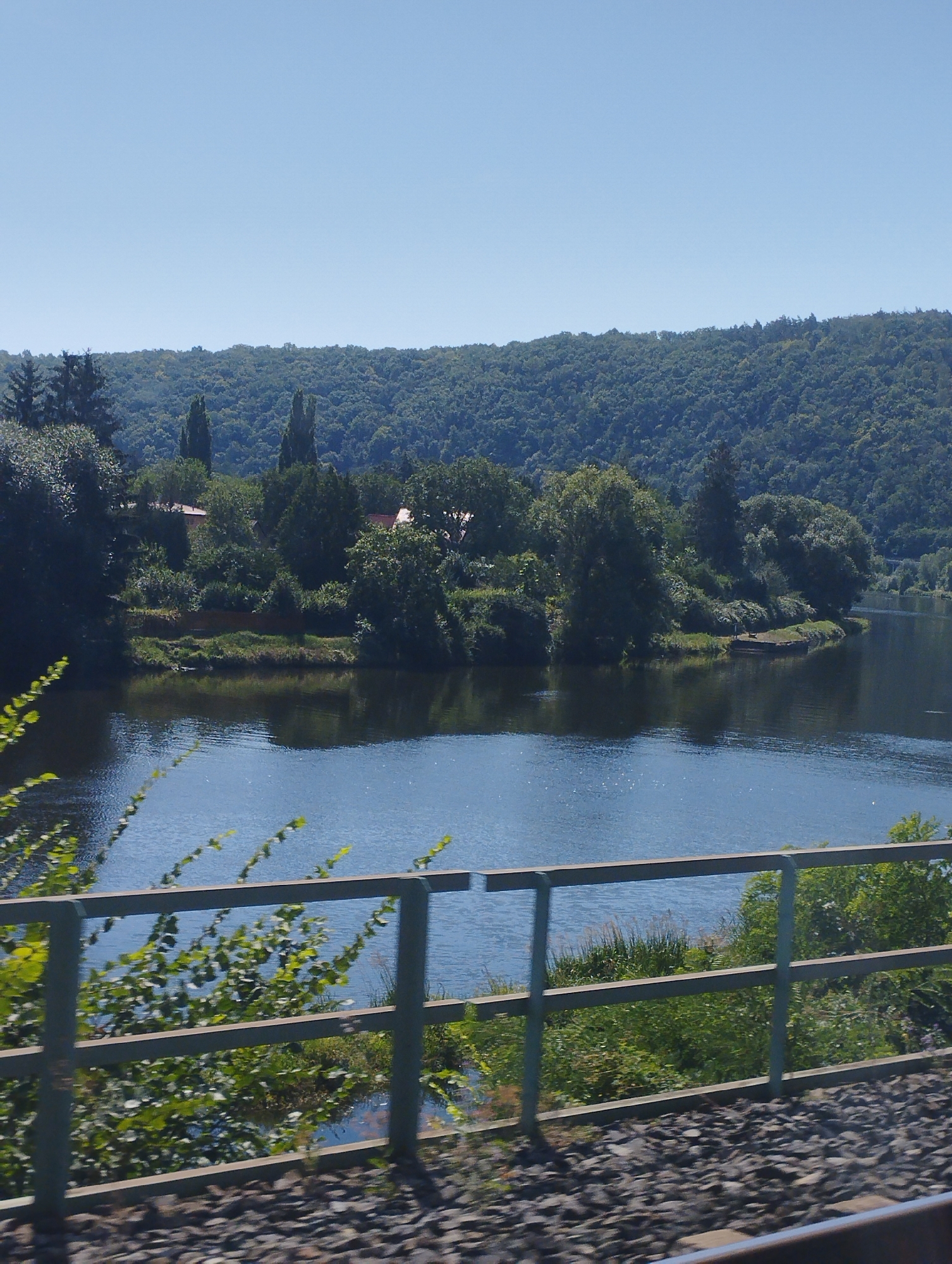 The image captures a serene riverside landscape under a clear, bright sky. In the foreground, a weathered metal railing, likely part of a bridge or observation deck, spans across the frame, with the textured gravel of a railway track visible below it. Beyond the railing, lush green foliage and trees create a vibrant border between the foreground and the water. The river itself is a deep, reflective blue, with gentle ripples disturbing its surface. On the opposite bank of the river, a dense forest covers rolling hills that recede into the distance. A few scattered red-roofed buildings are nestled amongst the trees, suggesting a small settlement or scattered residences. The overall impression is one of natural beauty and tranquility, with a touch of human presence integrated into the landscape.