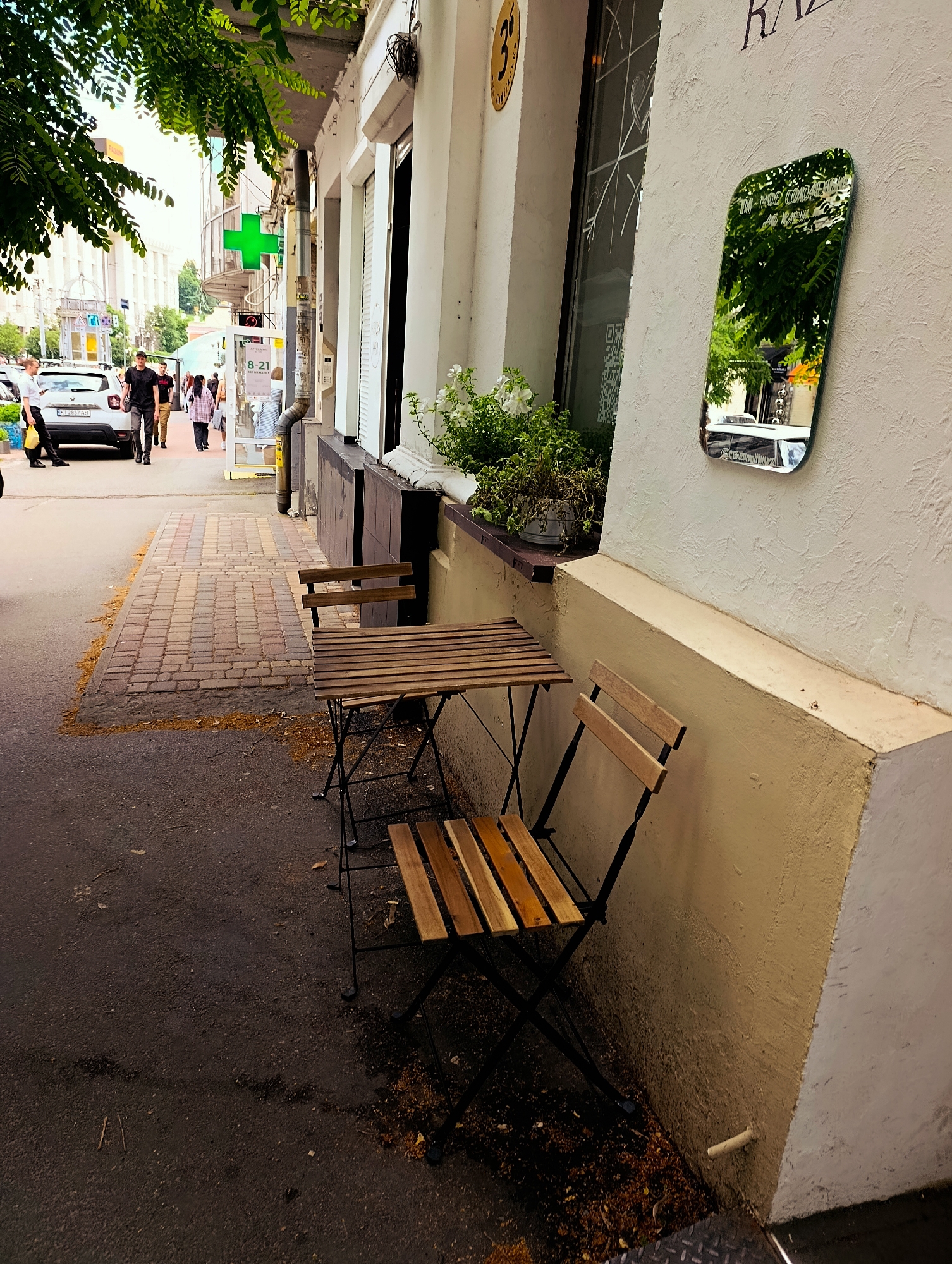 The image depicts a street scene in Kyiv, Ukraine, on a bright day. In the foreground, a small outdoor seating area with a wooden table and two chairs is set up on the pavement. The table and chairs are made of light-colored wood and have black metal frames, suggesting a casual café or restaurant setting.
Beyond the seating area, a brick sidewalk runs parallel to the street. Several people are walking along the sidewalk and on the street. One person, a man in a black t-shirt and jeans, is walking towards the camera. Another man in a white shirt and black pants is leaning against a white car parked on the street. A white car with a license plate "KI 2857 AB" is visible.
On the building to the right of the sidewalk, a distinctive green cross, commonly associated with pharmacies, is mounted. A sign beneath it reads "8-21". A small, rounded mirror is affixed to the building's exterior wall, reflecting the trees and sky above, as well as a portion of the street. The building itself is painted in light, neutral tones, with windows and architectural details characteristic of urban European architecture.
The scene appears to be a typical daytime street activity, with people going about their daily routines. The presence of the green cross and the text on the building suggest a commercial area, likely a pharmacy or a business with extended hours. The overall impression is one of an ordinary day in a bustling urban environment.