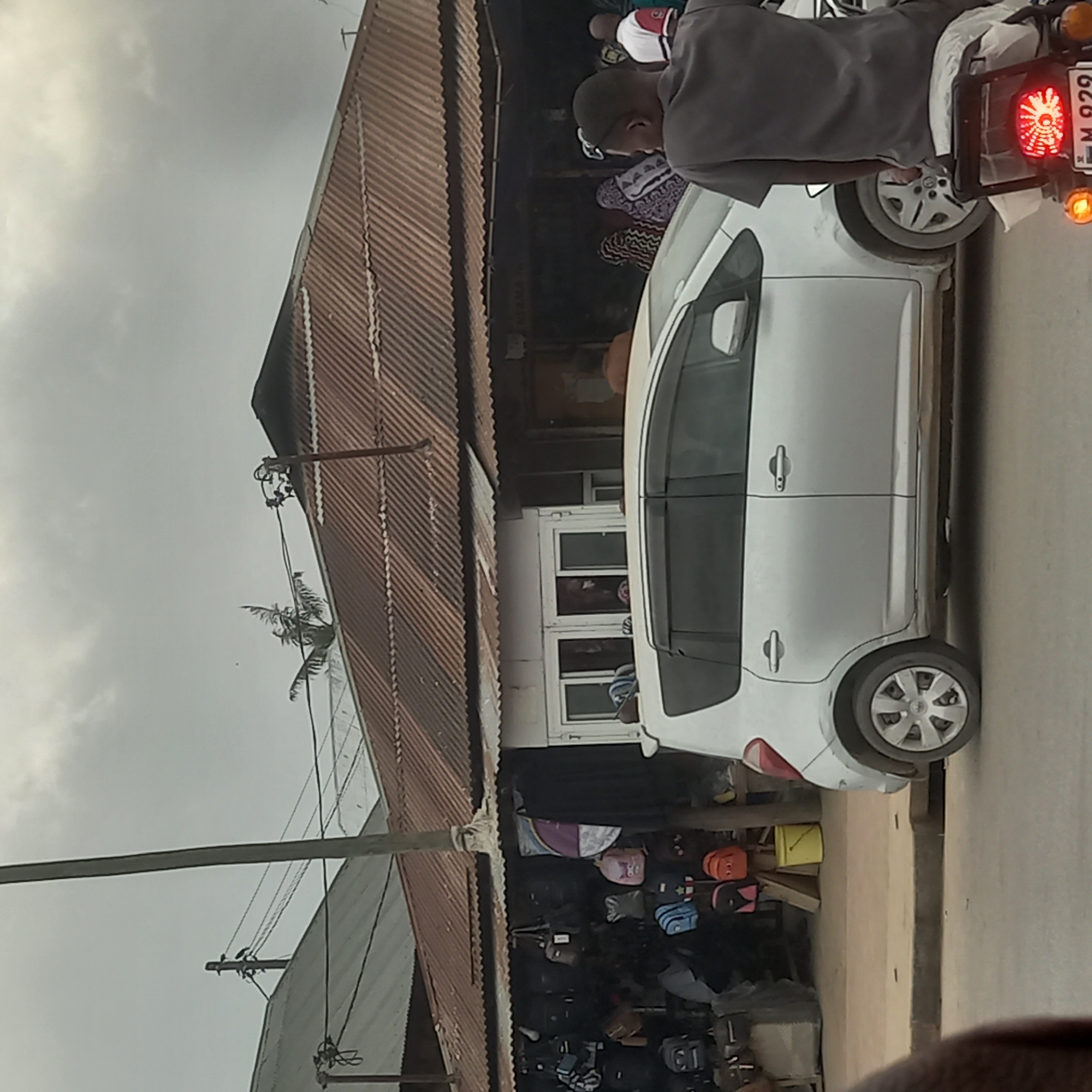 The image depicts a street scene in Bububu, Tanzania, with a focus on a small roadside market. A silver hatchback car is parked, partially obscuring a shopfront that displays various bags and backpacks. Behind the car, a man in a grey t-shirt is seated on a motorcycle, looking away from the camera. The market stall has a corrugated metal roof and is open to the street, with goods displayed both inside and outside. Several people, some wearing traditional clothing, are visible within the market area. Utility poles and overhead wires are present in the background, along with other buildings and a cloudy sky. The street appears to be paved.