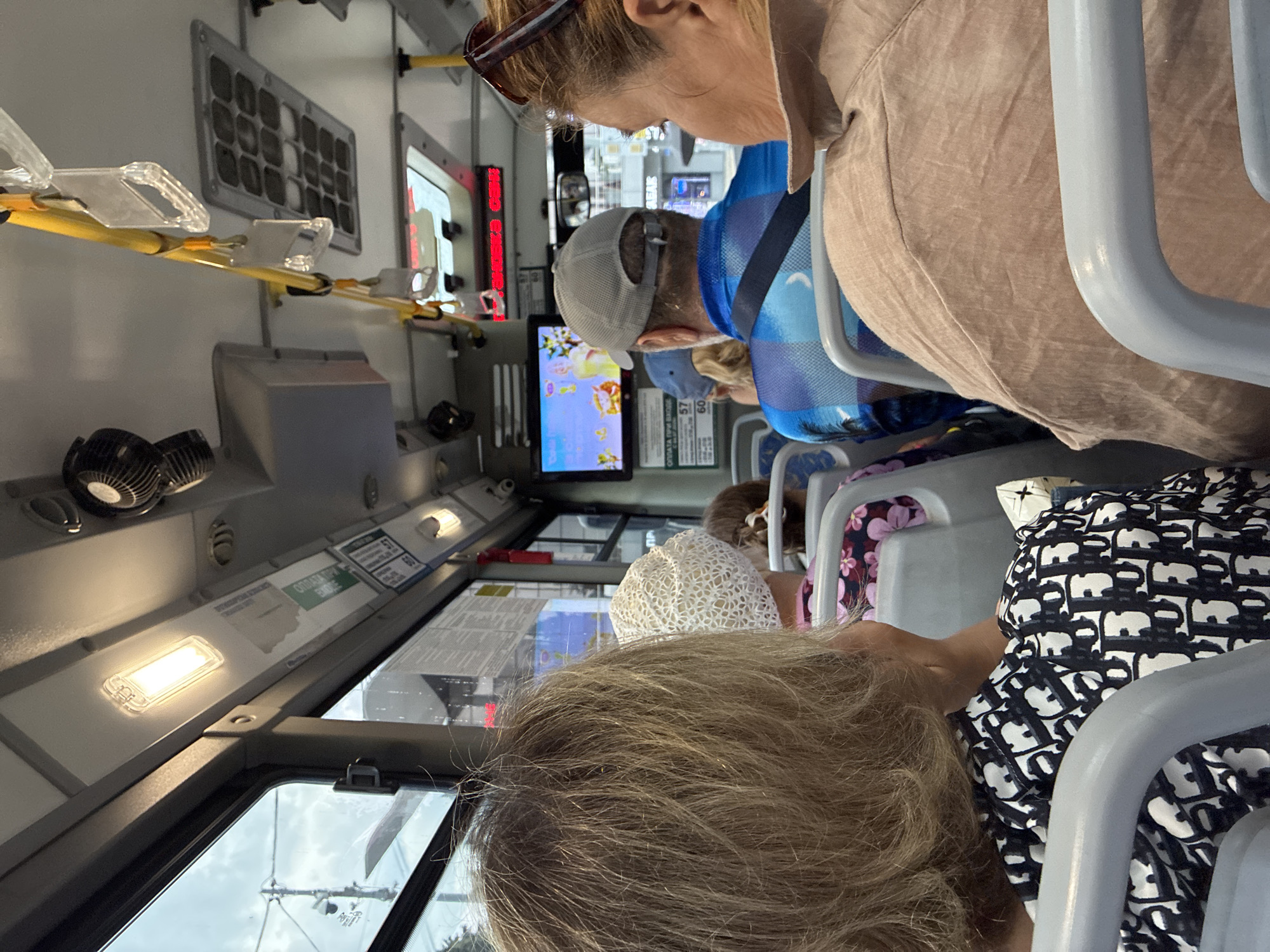 The image captures the interior of a bus in Sochi, Russia, during daylight hours. The perspective is from a seated passenger looking forward. The bus appears to be in motion, suggested by the slight blur of the exterior scene visible through the windows and the overall sense of being inside a moving vehicle.
In the foreground, the back of a woman's head with blonde, slightly messy hair is prominent on the left. She is wearing a black and white patterned top with a repeating 'Dior' logo. To her right, a woman in a white crocheted hat is partially visible. Further ahead, a man wearing a grey baseball cap and a blue checkered shirt is seated. On the right side of the frame, a woman with blonde hair, wearing sunglasses on her head and a tan-colored top, is looking towards the right.
The bus interior is equipped with standard features. Overhead, there are fluorescent lights, ventilation grilles, and yellow grab handles for standing passengers. A small screen is mounted on the ceiling in the middle, displaying a colorful cartoon. Signs and advertisements are affixed to the ceiling panels. The seating appears to be grey plastic with metal frames. The windows offer glimpses of trees and utility poles outside, consistent with an urban or suburban environment. The overall atmosphere is that of ordinary public transportation.