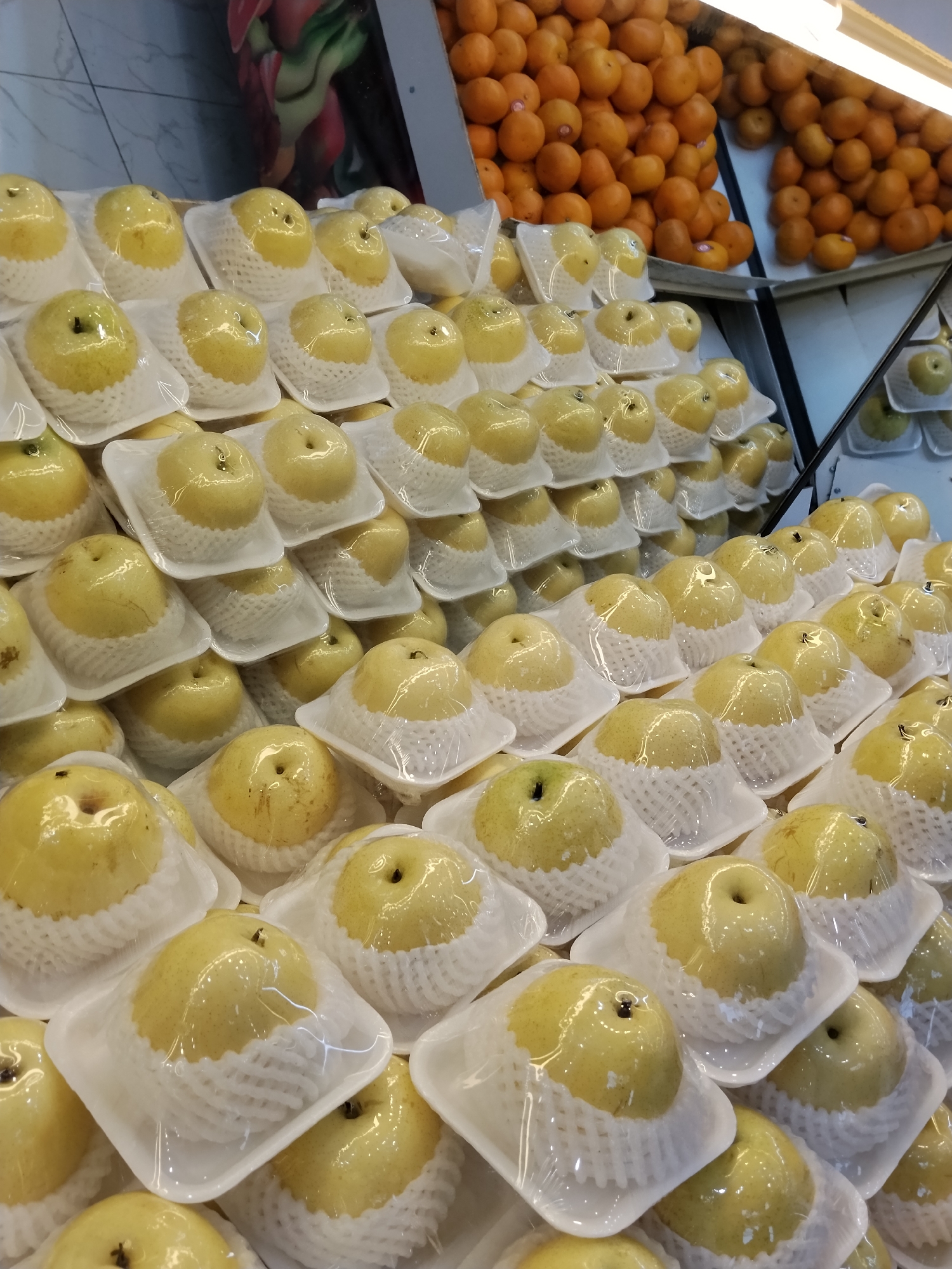 A close-up, eye-level shot captures an abundance of yellow-green pears, each individually wrapped in clear plastic and nestled in white foam padding. The pears are arranged in neat rows on white plastic trays, creating a visually organized display. In the background, a shelf displays a large pile of small, round, orange fruits, likely tangerines or oranges. The scene appears to be inside a market or grocery store, possibly in the city of Ciruas, Indonesia, given the context. The lighting suggests an indoor environment with artificial illumination from above. There are no people visible in the frame.