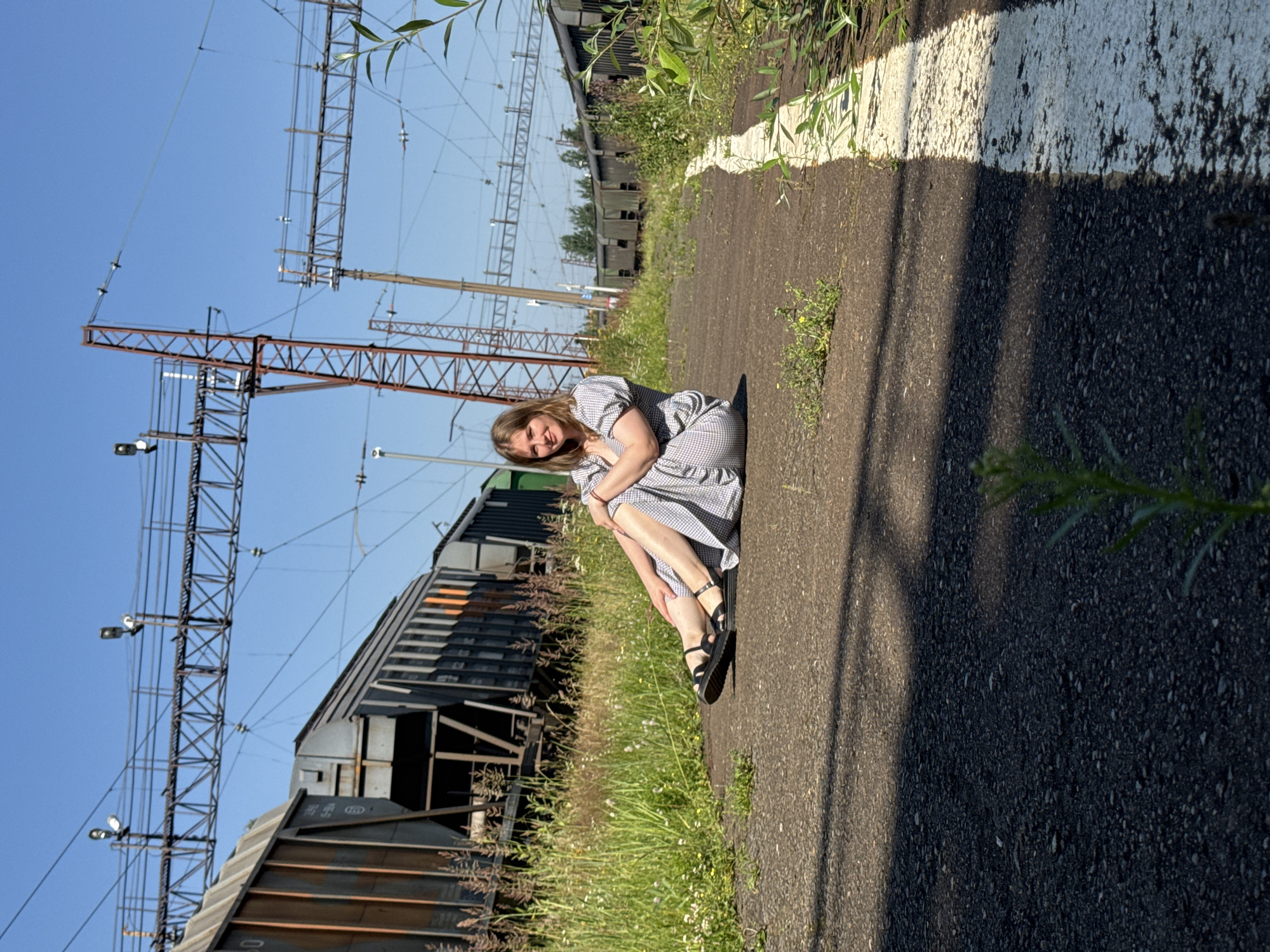 A young woman with blonde hair, wearing a grey and white checkered dress and black sandals, sits on the ground at a train station. She is positioned on the right side of the frame, with her legs crossed and her left arm resting on her bent knee. Behind her, a train with dark carriages is parked on the tracks, with overhead power lines and metal support structures visible against a clear blue sky. The ground is asphalt, with some patches of grass and weeds growing through it. A white line is painted on the asphalt to the right of the woman, marking the edge of the platform. The scene appears to be in an outdoor industrial setting.