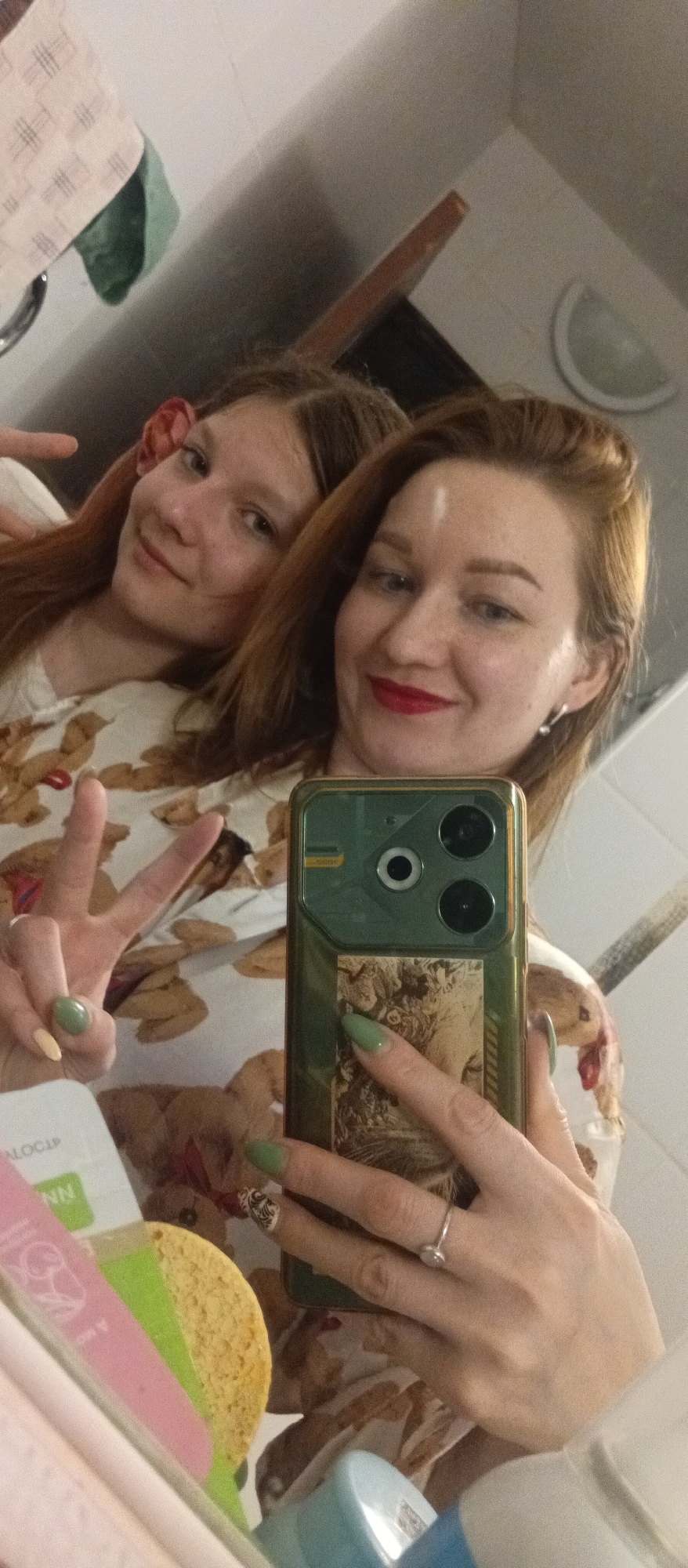 A mirrored selfie shows two light-skinned women, likely indoors in an unknown location in the city of Unknown, Unknown. The woman in the foreground holds an olive green cell phone with a vintage-style design on the back; the phone has multiple camera lenses. Her nails are painted pale green, except for one yellow one on the hand holding the phone, which also wears a silver ring. She wears red lipstick and dangling earrings. A younger woman, possibly a daughter, is behind and to the left, holding up a peace sign. Both women wear a white, patterned shirt. To the left of the phone are several objects including a sponge and product packaging; in the upper background there's a towel rack and a small shelf-like structure. The general setting appears to be a bathroom.