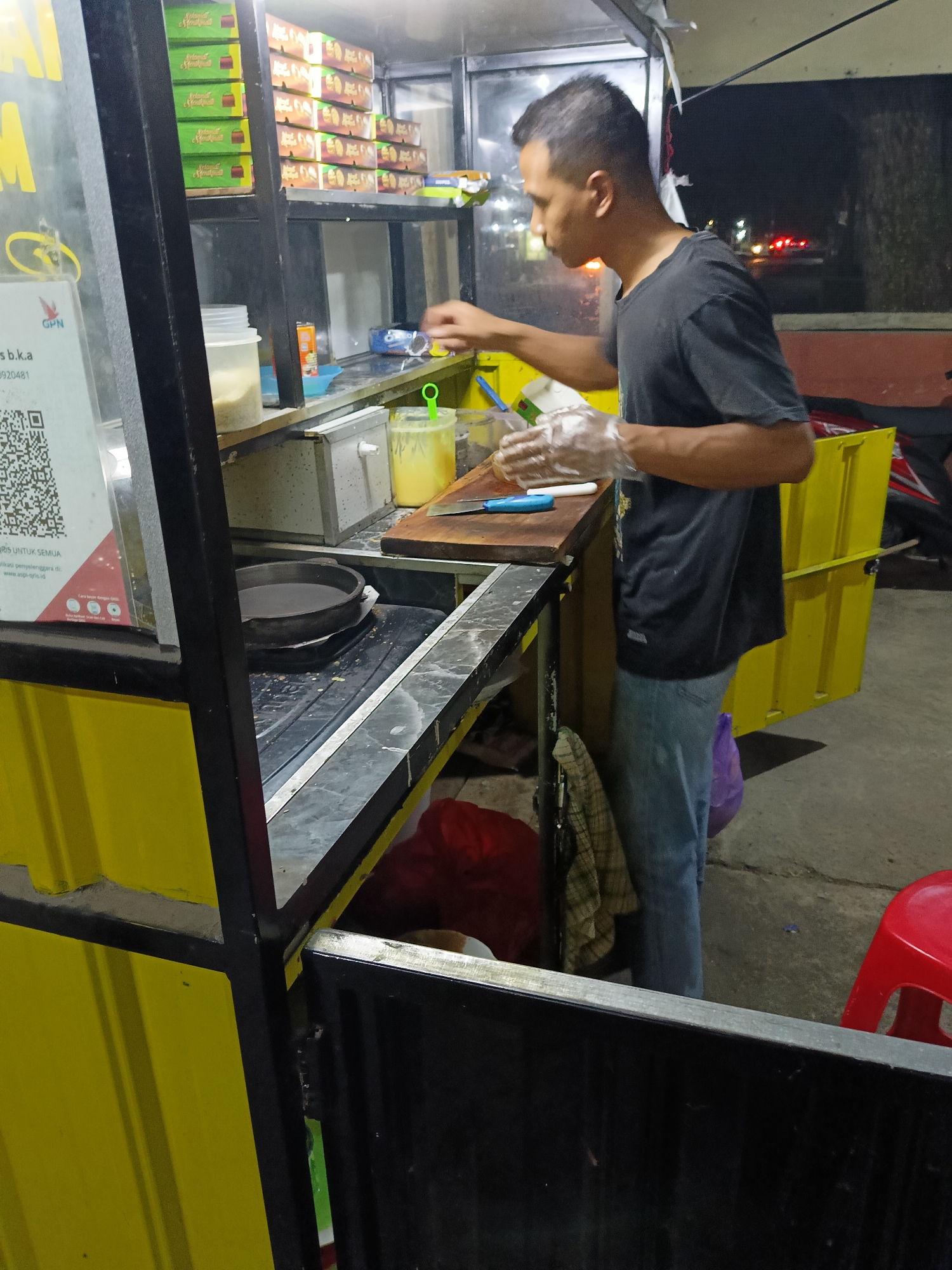 An Indonesian man, wearing a dark t-shirt and blue jeans, is preparing food at a street food stall at night in Manisrenggo, Indonesia. He is wearing clear plastic gloves and is holding a knife over a wooden cutting board. In front of him, there is a flat-top griddle with a dark substance in it. Behind him, shelves are stacked with boxes of packaged food items. The stall has bright yellow siding and is illuminated by artificial lights. A red stool is visible to the right of the frame. The background suggests a street scene with faint lights from passing vehicles.