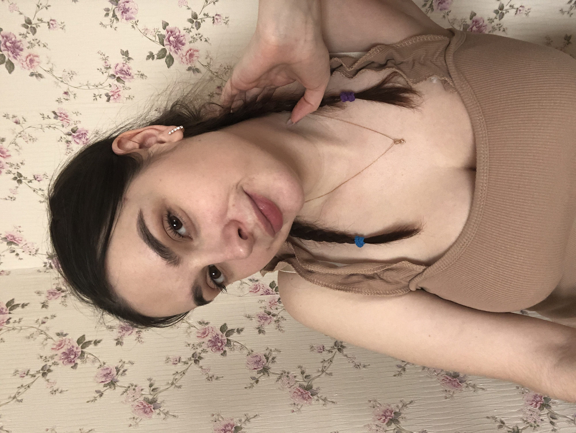 The image depicts a female individual indoors in Noviy, Russia. She is positioned from the chest up, looking directly at the camera with a slight smile. Her dark brown hair is styled with two braids, one on each side, secured with a blue elastic band on the viewer's left and a purple elastic band on the viewer's right. Her visible left ear bears an earring composed of multiple small, clear stones arranged in a curved line. A thin, gold-colored chain with a small pendant is visible around her neck. She is wearing a light brown, ribbed tank top with thin straps. Her right hand is raised, with fingers lightly touching the hair and neck area on the right side of her head. The background features light-colored wallpaper with a repeating pattern of pink and purple floral motifs and green foliage.
