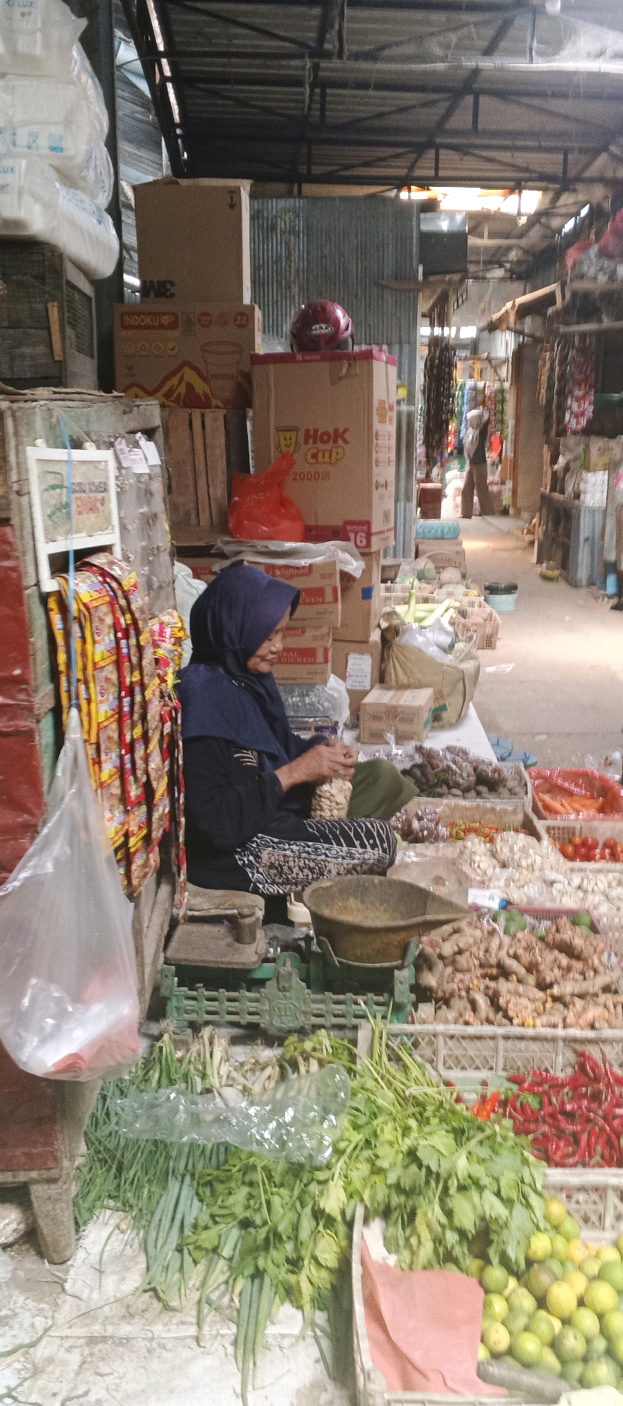 In a bustling outdoor market stall in Sekaran, Indonesia, a woman wearing a dark blue hijab and a black top is seated, sorting through a pile of white nuts. She is positioned behind a green weighing scale with a brown metal bowl. To her left, a red chair is partially visible, and on the scale, a bundle of what appears to be dried goods is being handled by the woman.
The stall is laden with a variety of produce. In the foreground, bundles of green onions and leafy vegetables are laid out. Behind these, several crates overflow with colorful items: bright red chilies, tomatoes, and other assorted produce. A large pile of ginger-like root vegetables is prominently displayed in a crate in the lower right portion of the image. To the left of the woman, a white board displays some items, and to her left are hanging packets of snacks or goods, predominantly yellow and red in color.
The background reveals the market's infrastructure. Stacks of cardboard boxes, some labeled with brand names like "3M" and "HoK Cup," are piled high. A red helmet rests on top of one of these boxes. The market appears to be covered by a corrugated metal roof supported by a metal frame. In the distance, the aisle of the market is visible, with other stalls and shoppers, one person walking away from the camera. The ground is a mix of concrete and tiled surfaces. The overall scene depicts a vibrant, busy market environment with fresh produce and various goods for sale.