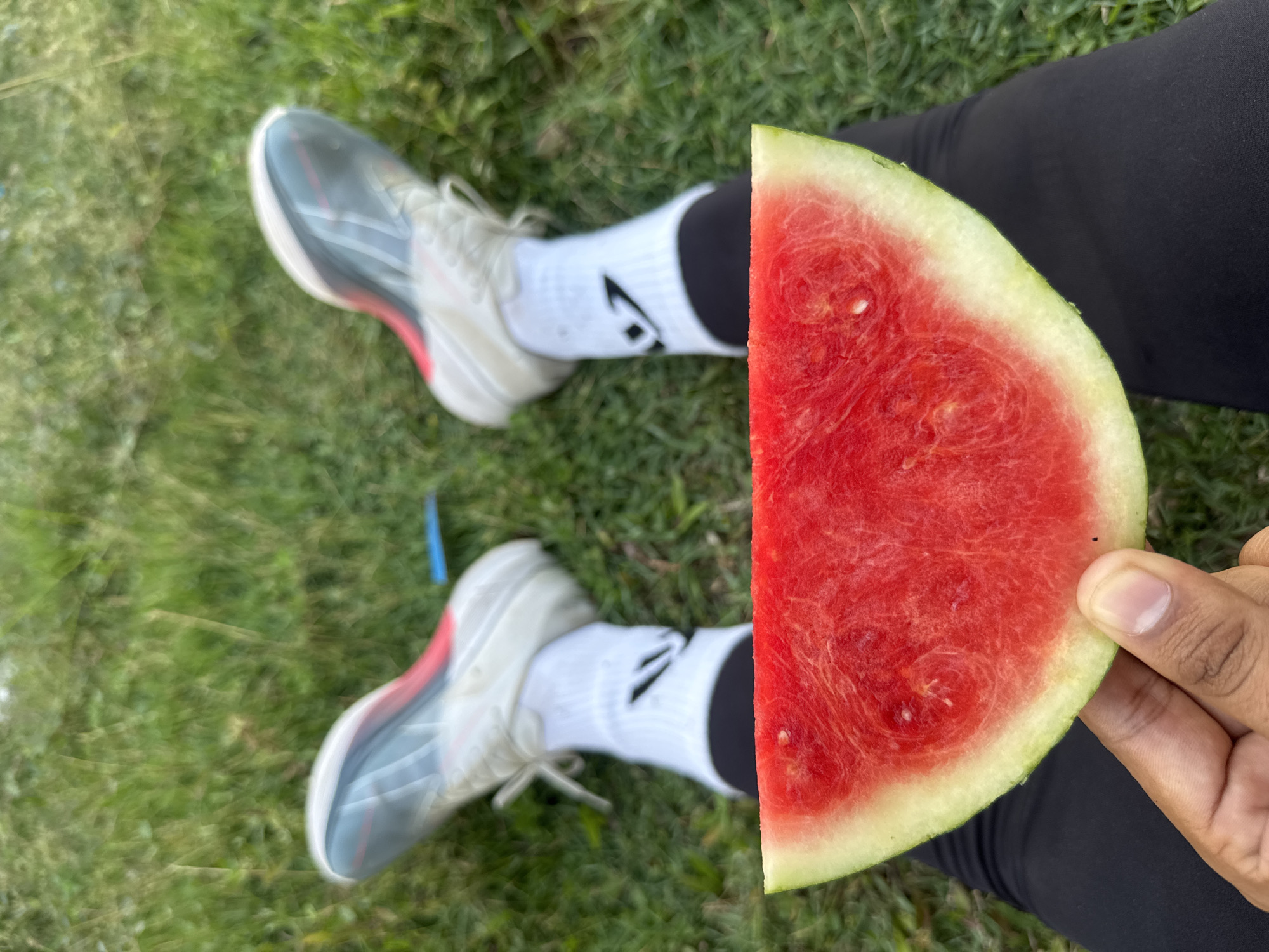 In Meureudu, Indonesia, a close-up, low-angle perspective shot captures a slice of watermelon being held by a person. The watermelon slice, with its bright red flesh and green rind, is positioned in the foreground. Behind it, the person's legs are clad in black leggings and white socks with a black logo. The person is wearing athletic shoes. The background consists of a blurred expanse of green grass, suggesting an outdoor setting.