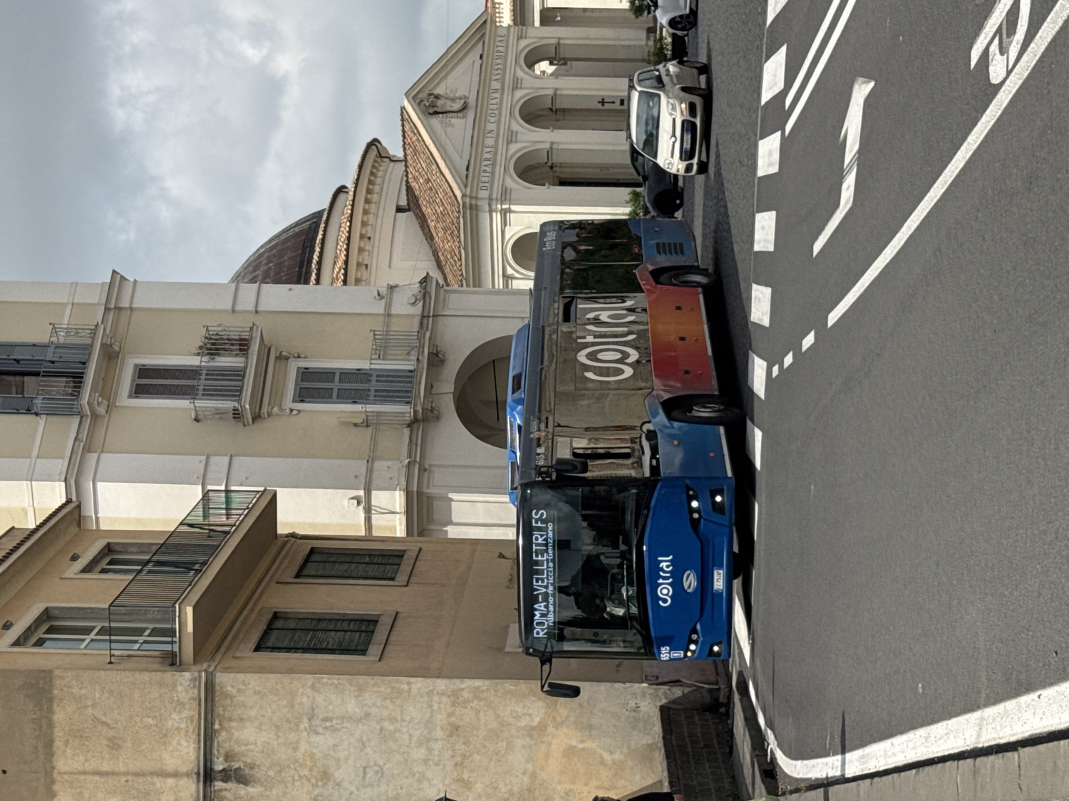 This daytime image, taken in Ariccia, Italy, depicts a street scene under an overcast sky. The primary subject is a blue public transit bus, part of the "Cotral" fleet, which is prominently displayed with its logo on the front and side. The bus is marked with "ROMA-VELLETRI FS" as its destination, with intermediate stops "Albano-Ariccia-Genzano" listed below. The bus identification number "6515" is visible on the front. Behind this bus, another "Cotral" bus, partially visible, is painted in an orange and white livery and also bears the "Cotral" and "Eco Bus" branding.
The buses are positioned on an asphalt road featuring white painted markings, including directional arrows and dashed lines indicating traffic lanes. Further down the street, a white car and another vehicle are visible.
To the left of the road, several buildings of varying architectural styles are present. In the foreground, a building with multiple windows, some fitted with closed shutters and external metal grates, is seen. In the background, a large, ornate building, likely a church or a significant public structure, features a prominent dome and a classical facade. Text is visible on this facade, reading "DEIPARAE IN COELVM ASSYMPTAE." The scene captures a blend of modern public transportation against a backdrop of historic Italian architecture.