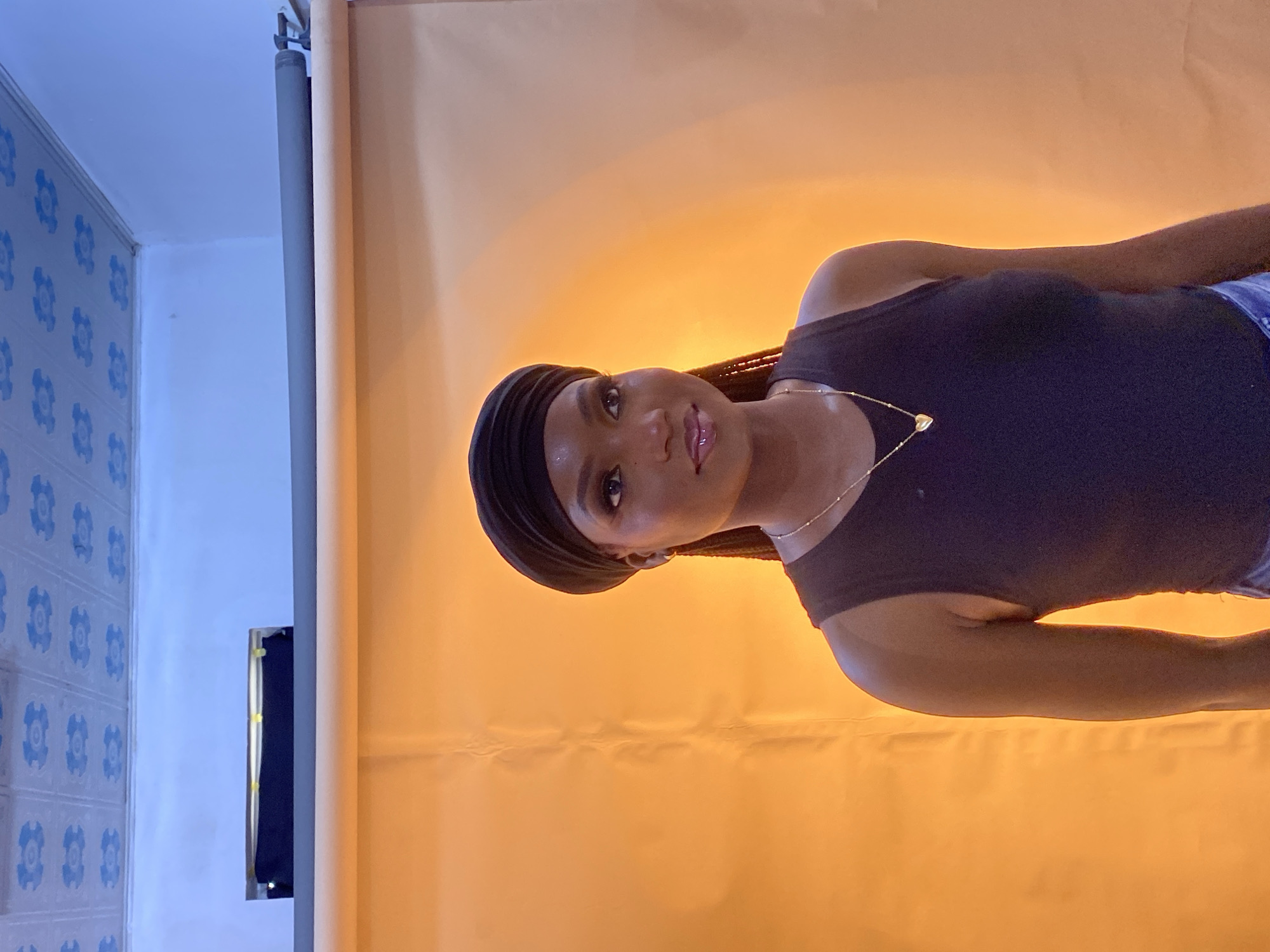 A young Black woman is posing in front of an orange backdrop. She is wearing a black tank top and a black headscarf. A thin gold chain with a heart-shaped pendant rests on her chest. Her hair is braided and extends down her back. The background appears to be a studio setting, with a rolled-up backdrop and a ceiling with blue floral patterns. The lighting is warm and casts a glow on the woman and the backdrop. There are no observable elements that suggest a location in Gura Top, Nigeria.