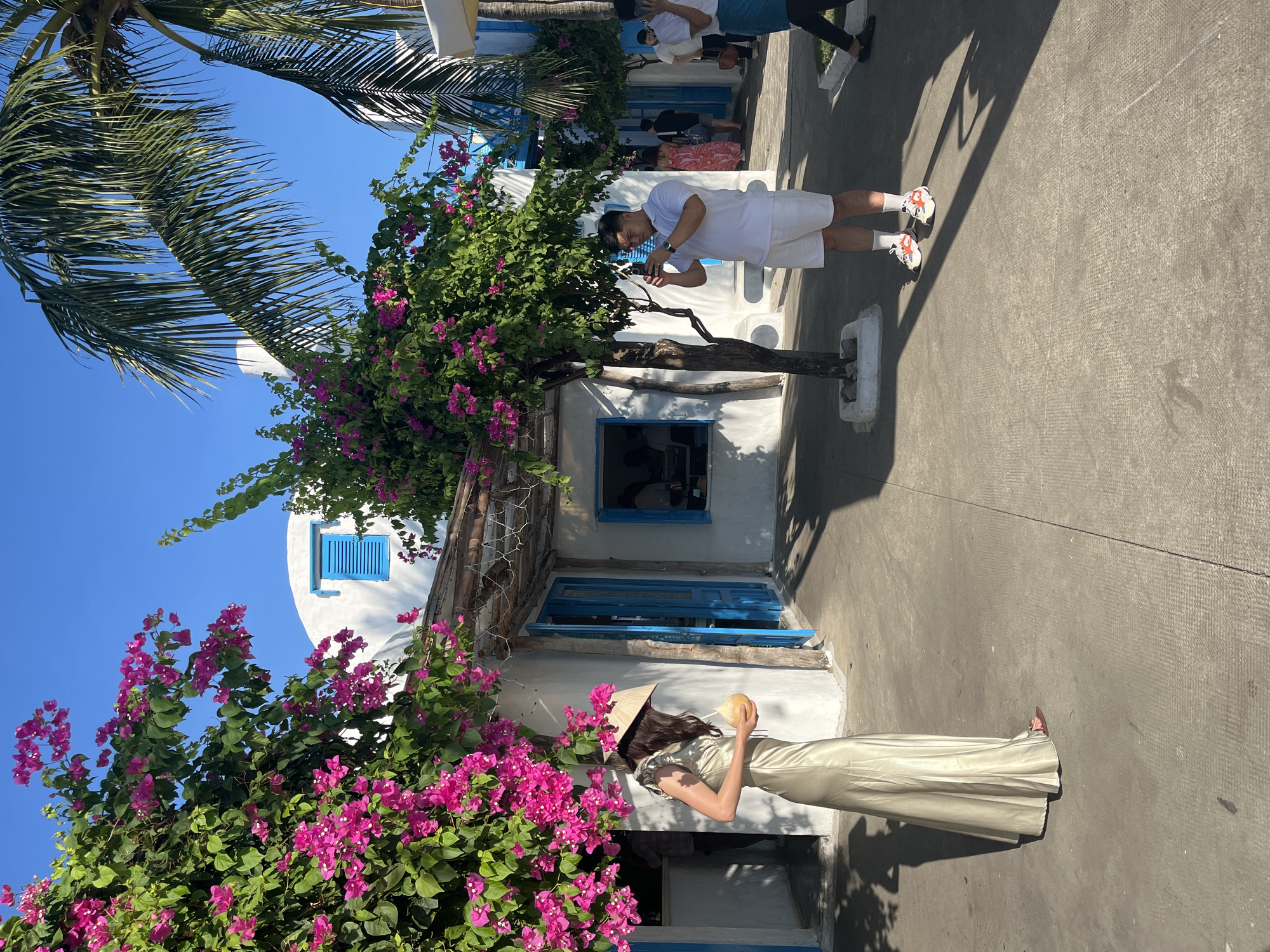 The image depicts a scene in Đà Nẵng, Vietnam. A woman in a light-colored gown and a conical Asian hat holds a yellow fruit or drink near a whitewashed building adorned with blue trim and vibrant pink bougainvillea. A man in white clothing is looking at his phone by a tree in a similar building style. Other people are visible in the background. The day is sunny, and shadows are cast on the concrete ground. Palm fronds are visible above.