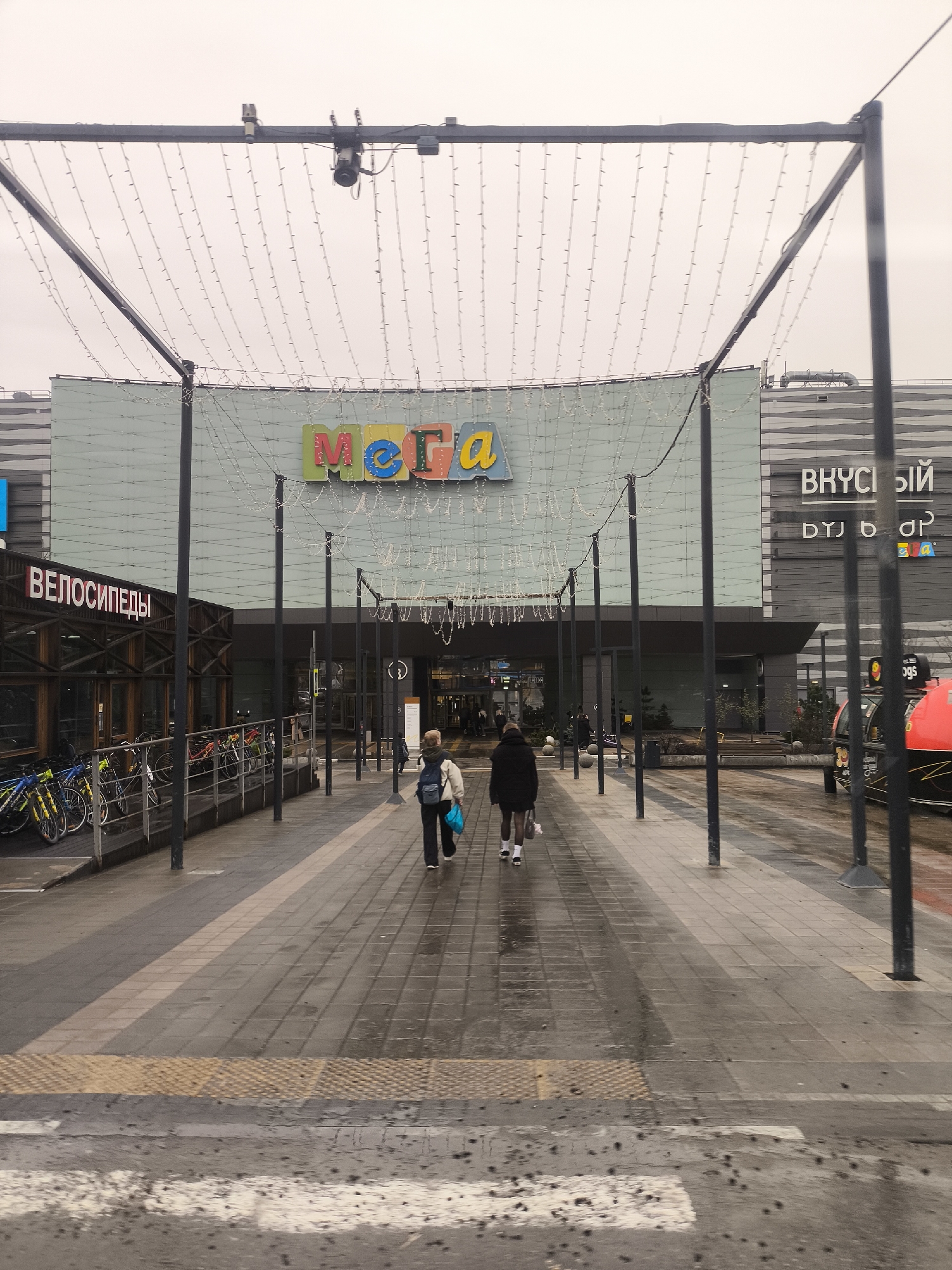 The image depicts the entrance to the "MEGA" shopping center in Khimki, Russia. Two people are walking towards the entrance. The area is paved with wet tiles. The sky is overcast, suggesting a rainy or recently rainy day. The MEGA sign is visible above the entrance. To the left, there is a building with the word "ВЕЛОСИПЕДЫ" (BICYCLES) in Russian, with bicycles parked nearby. To the right, part of a sign with "ВКУСНЫЙ" (TASTY) in Russian can be seen. Black metal beams and strung lights frame the entrance. A security camera is mounted on one of the beams. A pedestrian crosswalk is visible in the foreground. A red taxi can be seen on the right.