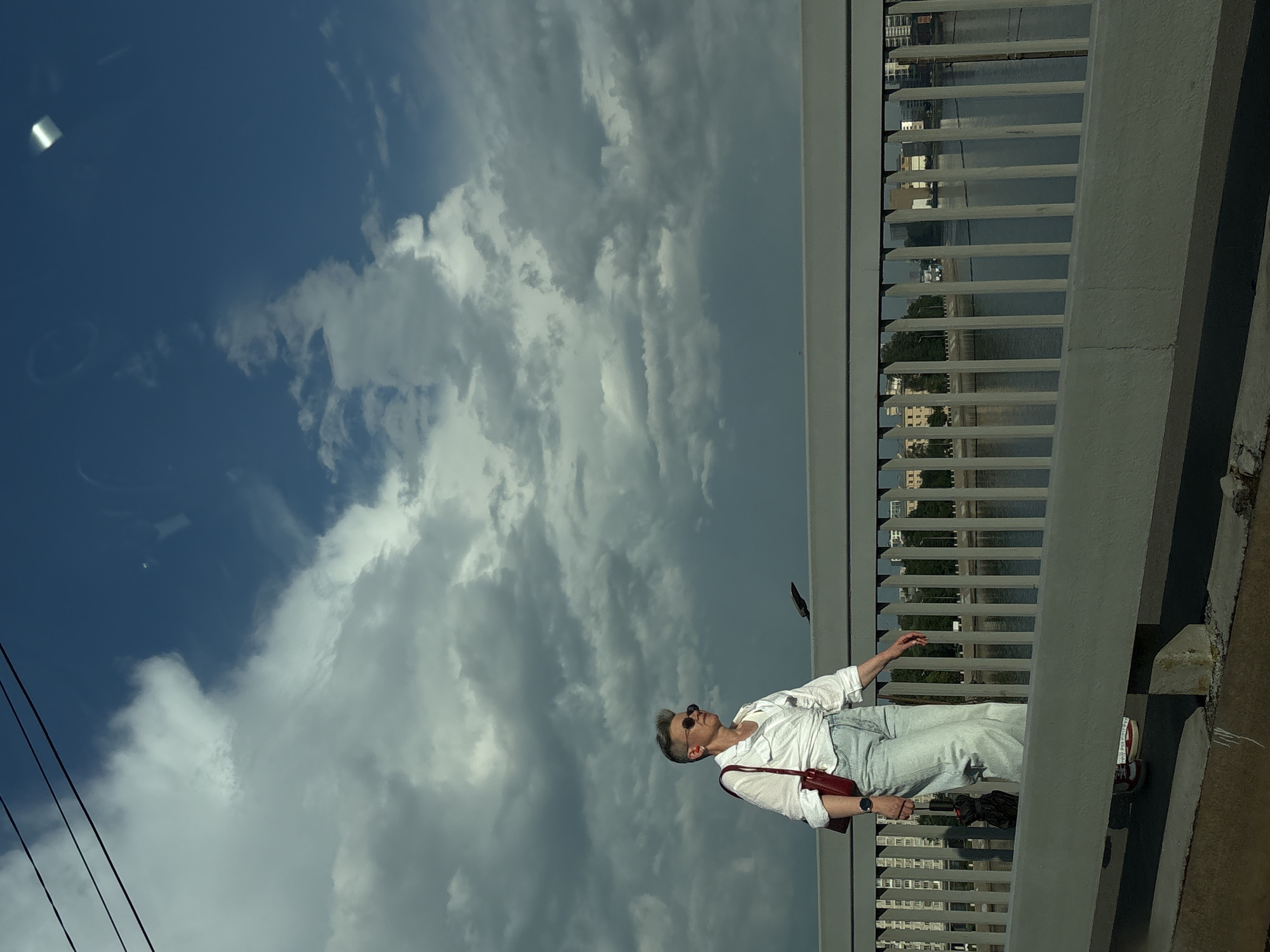 A woman with short gray hair and sunglasses walks across a bridge in Saint Petersburg, Russia, on a cloudy day. She is wearing a white shirt and light blue jeans and carries a red handbag and a black umbrella. Buildings and trees line the riverbank in the background. The sky is filled with dramatic clouds.