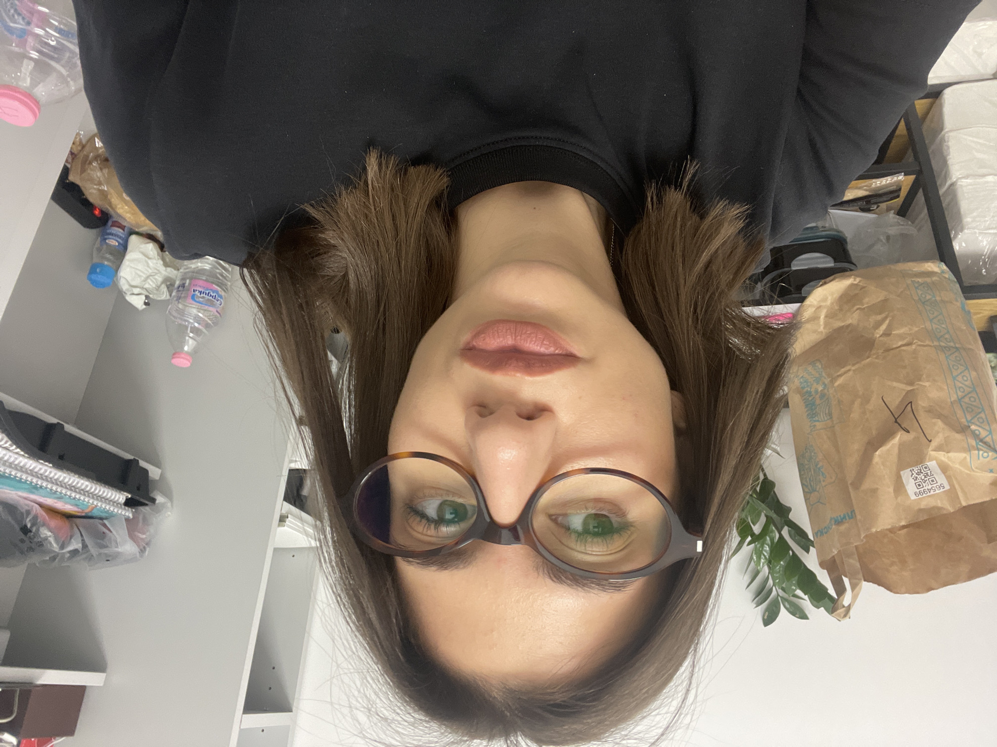 A woman with brown hair and glasses is facing the camera. She is wearing a black long-sleeved shirt. Her lips are slightly parted.
In the background, a white wall is visible. A shelf with various items is to her right, including a brown paper bag, boxes, and papers. To her left is a white cabinet with a stack of notebooks and papers.
A water bottle labeled "Gorna Banya" is visible. Gorna Banya is a neighborhood in Sofia, Bulgaria.