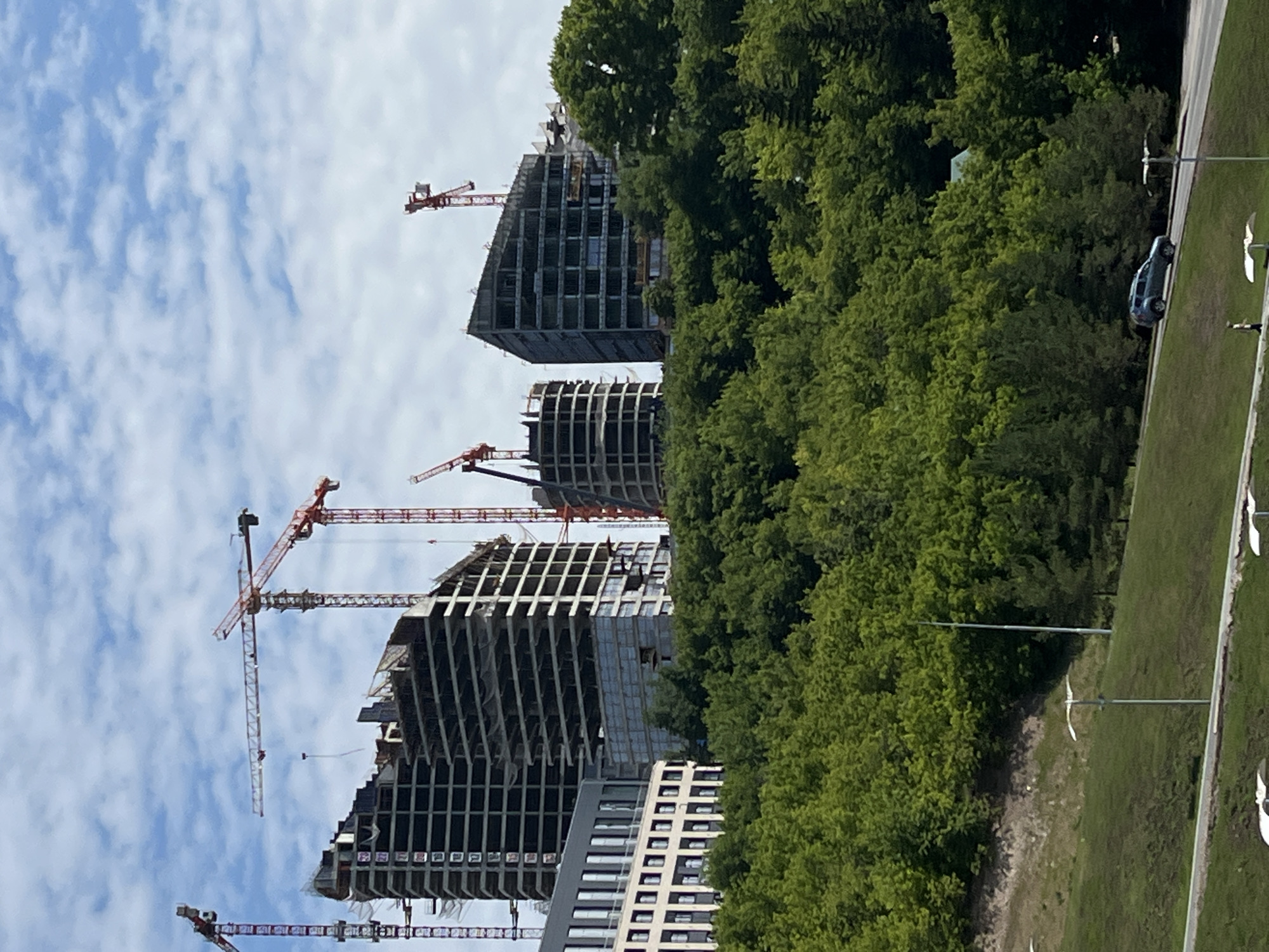 A construction site is depicted against a cloudy blue sky, featuring several unfinished high-rise buildings with visible scaffolding and cranes. The buildings are in various stages of completion, with some showing exposed concrete frames and others partially clad in glass. Cranes, primarily red, are positioned around the structures. Below the construction site, a dense, lush green forest covers a hillside. In the foreground, a grassy slope leads down to a paved path where a dark-colored SUV is driving uphill. There are also streetlights and what appear to be decorative lamp posts visible along the path and on the slope. The scene suggests ongoing urban development in a hilly, vegetated area.