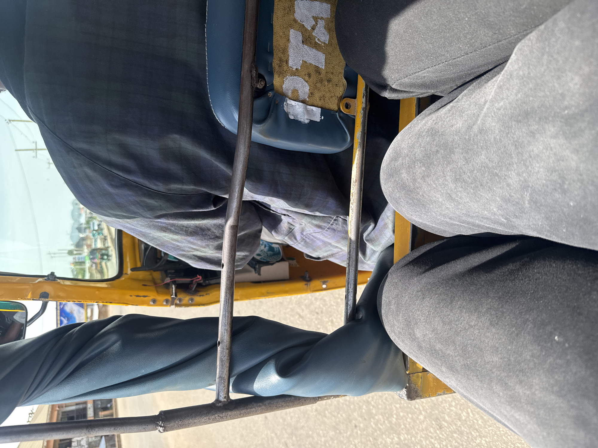 The image depicts the interior of a yellow auto-rickshaw, likely in Barakin Lerawi, Nigeria, viewed from the perspective of a passenger. The foreground is dominated by the legs of two individuals, clad in dark gray and black trousers, positioned on the left and right sides of the frame respectively. In the middle ground, a portion of the auto-rickshaw's structure is visible, including a metal bar and a blue seat. The seat has a yellow fabric element with white lettering, partially obscured and appearing to spell out "PTA". To the left, a twisted piece of blue upholstery is prominent. In the background, the driver, wearing a checkered shirt, is seated at the controls. The environment outside the auto-rickshaw is blurred but suggests a road with other vehicles and buildings, indicating an urban or semi-urban setting. The lighting suggests it is daytime, with sunlight casting shadows.