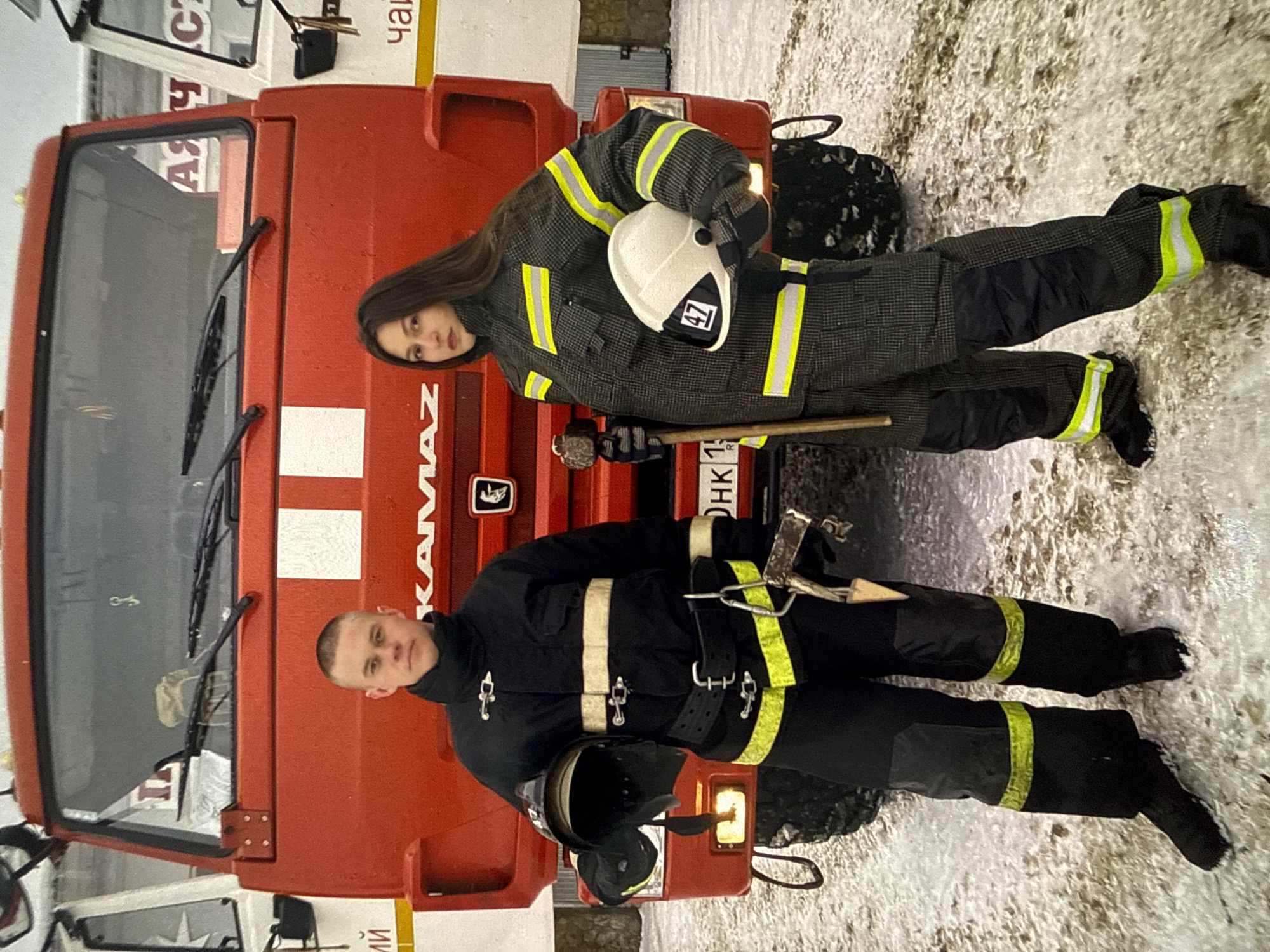 Two firefighters, a man and a woman, stand in front of a red KAMAZ fire truck. The man is on the left, wearing a black firefighter suit with yellow reflective stripes. He is holding a black helmet in his left hand and a tool, possibly an axe or a crowbar, in his right hand. The woman is on the right, wearing a dark grey patterned firefighter suit with yellow reflective stripes. She is holding a white helmet with a black and white emblem featuring the number "47" in her right hand. The ground is covered in snow and dirt. The fire truck has a large red front and "KAMAZ" written on it. Text is visible on the side of the truck, including what appears to be "АЯ СТ" and possibly Cyrillic characters related to the city of Chaikovsky, Russia. The overall scene suggests a training exercise or a response in a cold, outdoor environment.