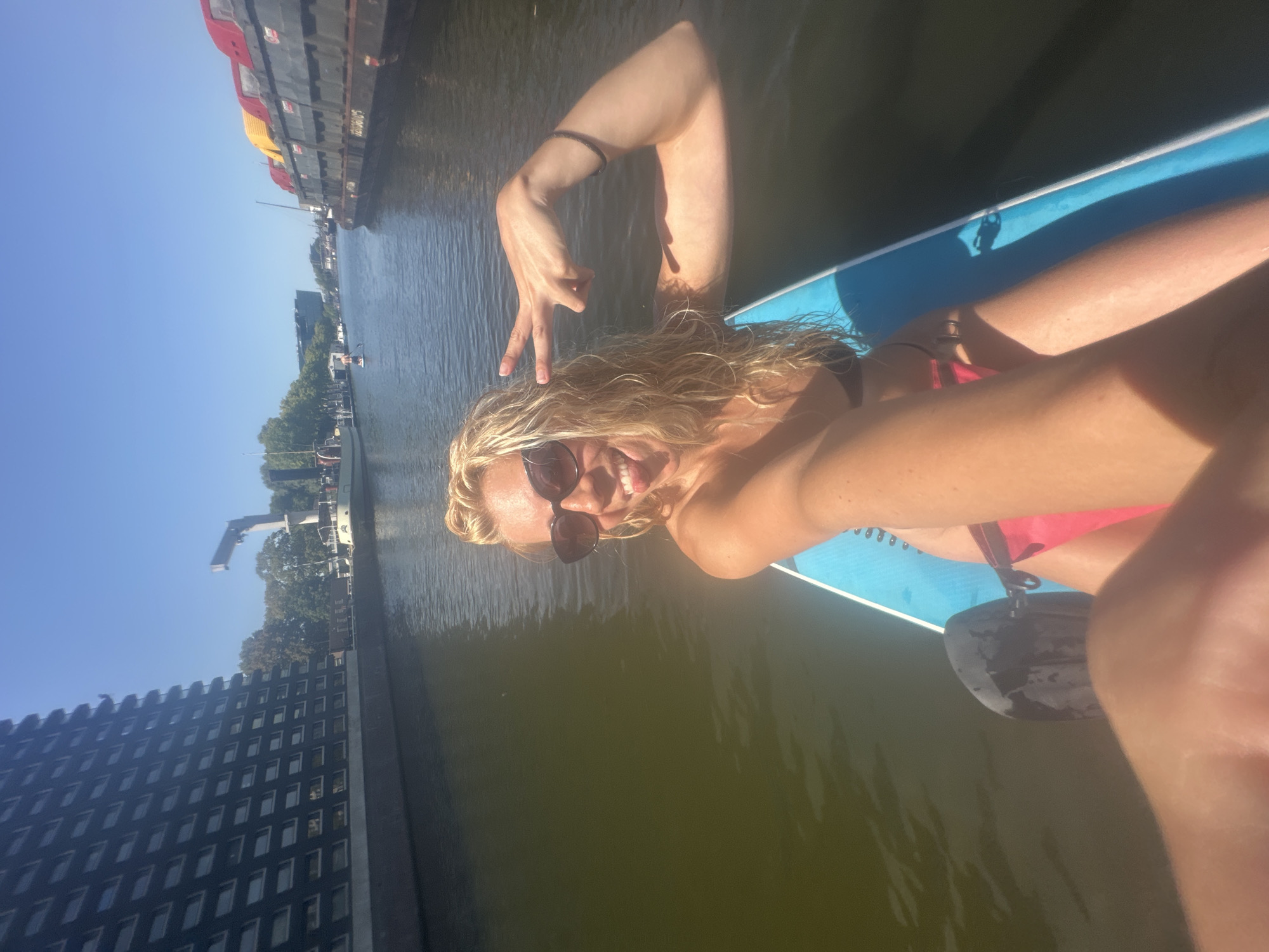 An outdoor, slightly low-angle, eye-level, medium shot captures a blonde woman paddleboarding on a sunny day in a canal in Amsterdam, Netherlands. She is wearing sunglasses, a black bikini top, and pink bottoms. She is sticking her tongue out and making a peace sign with her right hand. The paddleboard is blue and white, and the paddle is black. In the background, on the left, is a tall, dark building with many windows. On the right, there are large barges with red and yellow tops. The water is dark and reflects the sky and the surrounding structures. The sky is clear and blue.