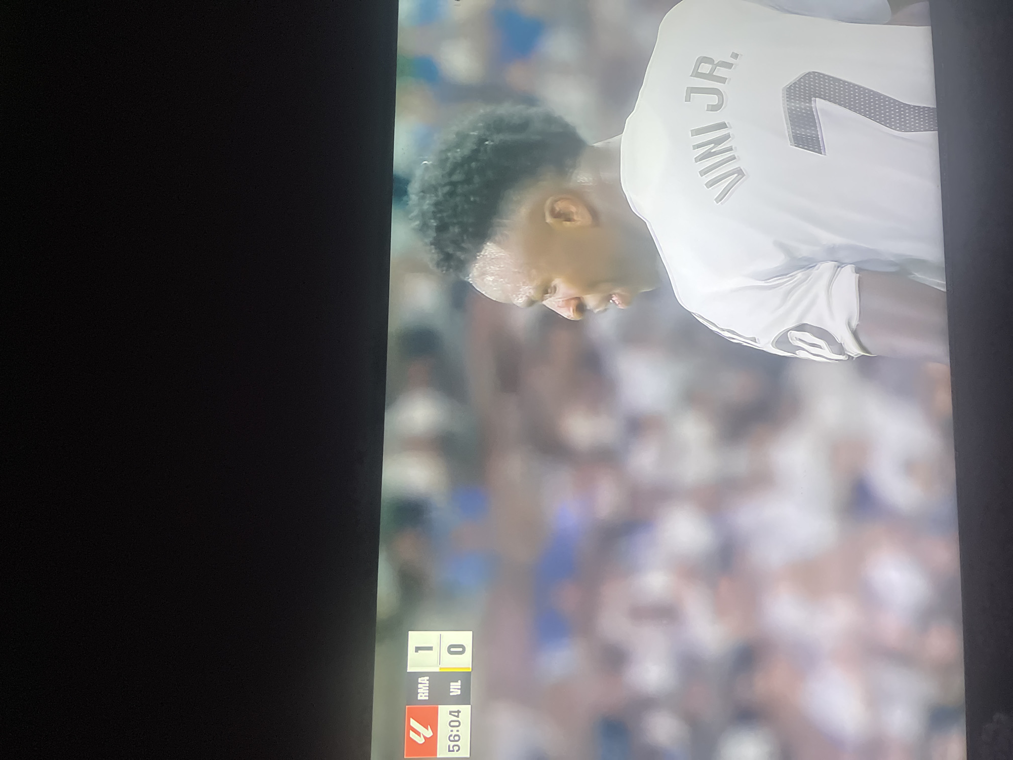 The image displays a portion of a television screen showing a football match. In the foreground, a player from Real Madrid, identified by the name "VINI JR." and the number "7" on his white jersey, is partially visible from behind. His hair is dark and curly, and he appears to be sweating. The background is blurred, showing a crowd of spectators and elements of a football stadium. In the upper left corner of the screen, a score graphic indicates "RMA 1" and "VIL 0", with a timestamp of "56:04". The city of Ruse, Bulgaria, is not depicted in this image.