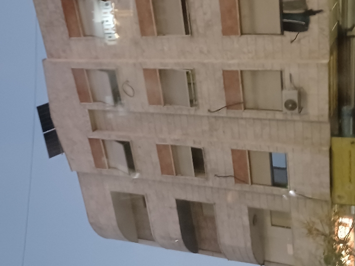 The image depicts the exterior of a multi-story building in Kafr Sousa Municipality, Syria, viewed from a slightly upward angle against a twilight sky. The building features a light-colored, possibly stone or concrete facade with a rounded corner on its left side.
On the roof, two dark, rectangular solar panels are visible. The building has multiple vertical rows of windows, most equipped with roll-down shutters. Several shutters are partially raised, revealing interior spaces or windows behind them. Specifically, shutters on the upper-left, middle-left (second from left), and upper-right windows are partially open. One window on the middle-left, beneath a partially raised shutter, displays a blue-tinted pane. A reflection of light-colored text, possibly Arabic characters, is visible on a window pane on the upper-right side.
Two white, rectangular outdoor air conditioning units are mounted on the facade. One is located on the lowest visible floor, second from the right, beneath a window. Another is situated on the far right of the lowest visible floor, below which dark fabric items, possibly laundry, are hanging. Dark, insulated electrical wires run vertically down the facade in several sections. A circular marking is present on the facade between windows on the middle floor, second from the left. A small, downward-pointing light fixture is visible on the middle-left section.
Below the main facade, a blurred yellow sign is partially visible at ground level, suggesting a commercial establishment. Blurred tree branches or foliage are also visible on the lower-left. The sky above is a clear, light blue to grey, indicative of dawn or dusk. No individuals are visible in the scene.