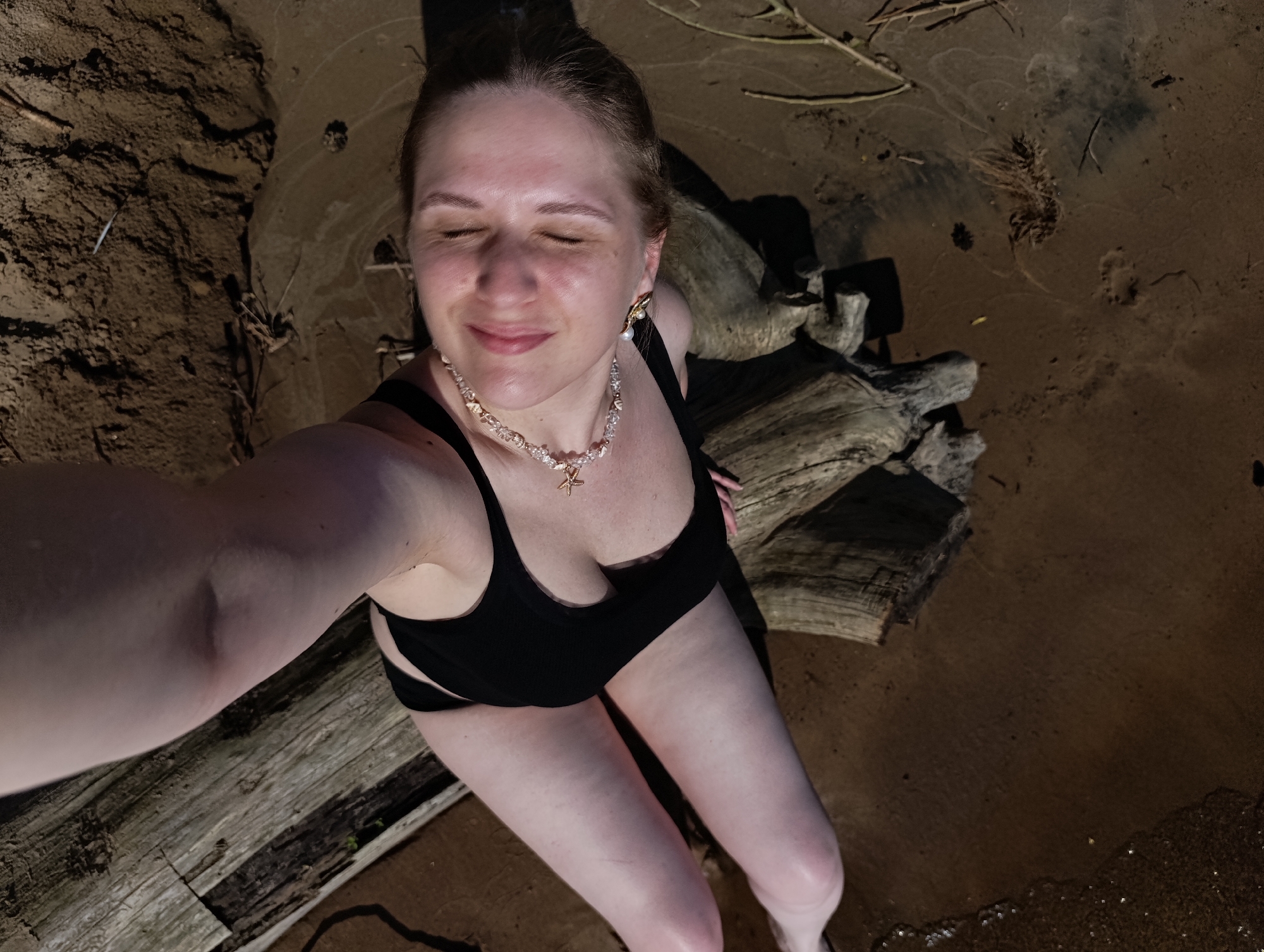 A woman is taking a selfie while sitting on a log near the water. She is wearing a black bikini and a necklace. The woman has her eyes closed and a smile on her face. The background is a sandy beach with some vegetation. The lighting suggests it is dusk or dawn.
