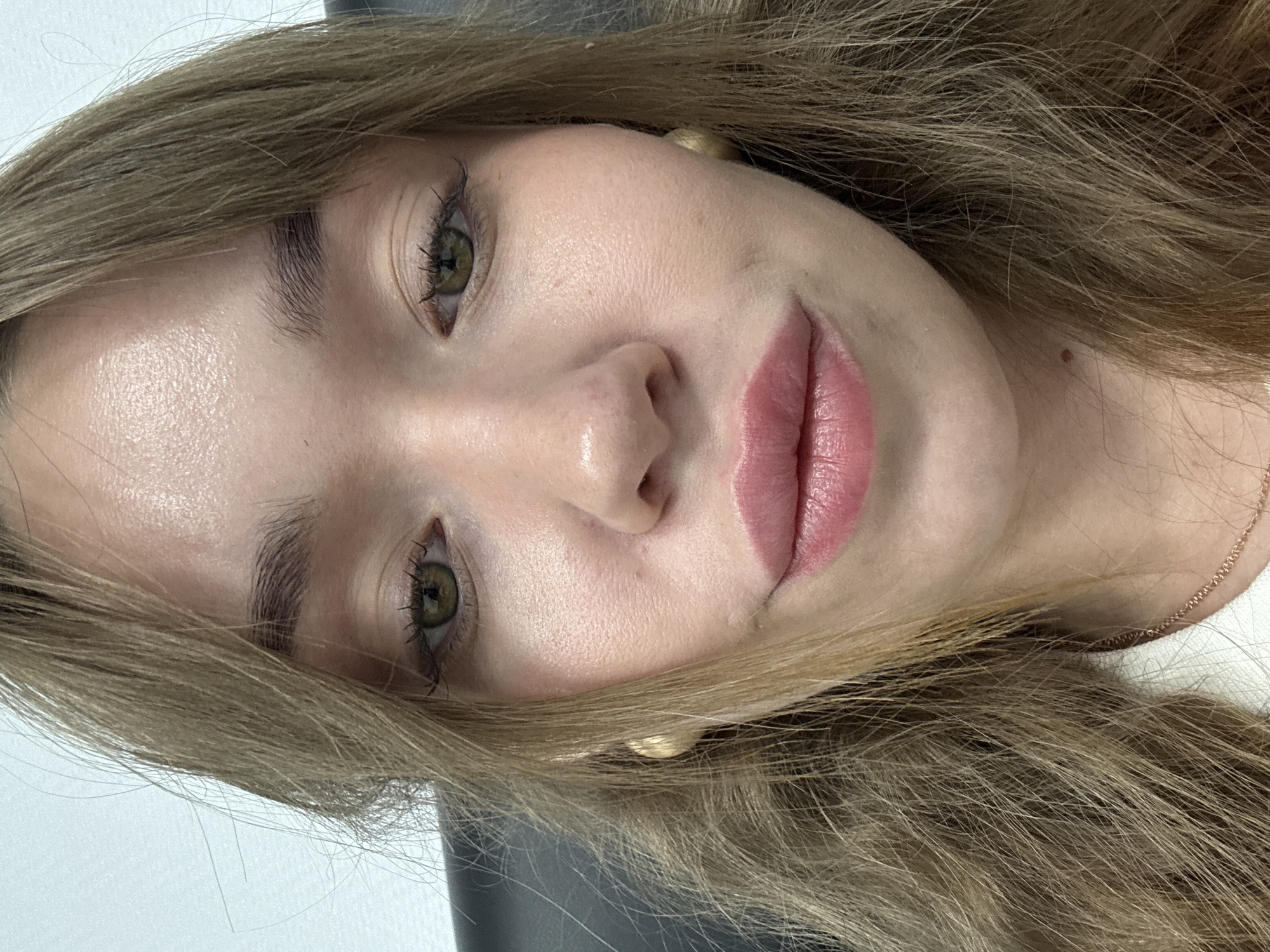 A close-up portrait of a young woman's face, rotated 90 degrees counter-clockwise, captures her looking directly into the camera. She has long, wavy hair, a blend of light brown and blonde hues, which frames her face and drapes over her shoulders. Her eyes are green or hazel, accentuated by dark mascara on her eyelashes, and her eyebrows are neatly shaped and full. Her lips are full and painted with a soft, natural pink lipstick.
The woman's expression is calm and neutral, with a hint of a gentle smile. Her fair skin appears smooth, with natural texture visible and a subtle sheen on her nose and forehead, suggesting either natural skin oils or subtle highlighting. She wears small, round, gold-colored stud earrings, one visible near her right ear (left side of the frame due to rotation), and a delicate, thin gold-colored chain around her neck, visible on the right side of the frame. A small, dark beauty mark is present on her upper neck/lower jawline area, visible on the right side of her face.
The background is softly blurred, showing a light, possibly white surface on the left and a darker, indistinct shape on the right, suggesting an indoor setting. There is no visible text in the image. No specific time of day or external weather conditions can be discerned from this close-up. The location context is Chaikovsky, Russia.