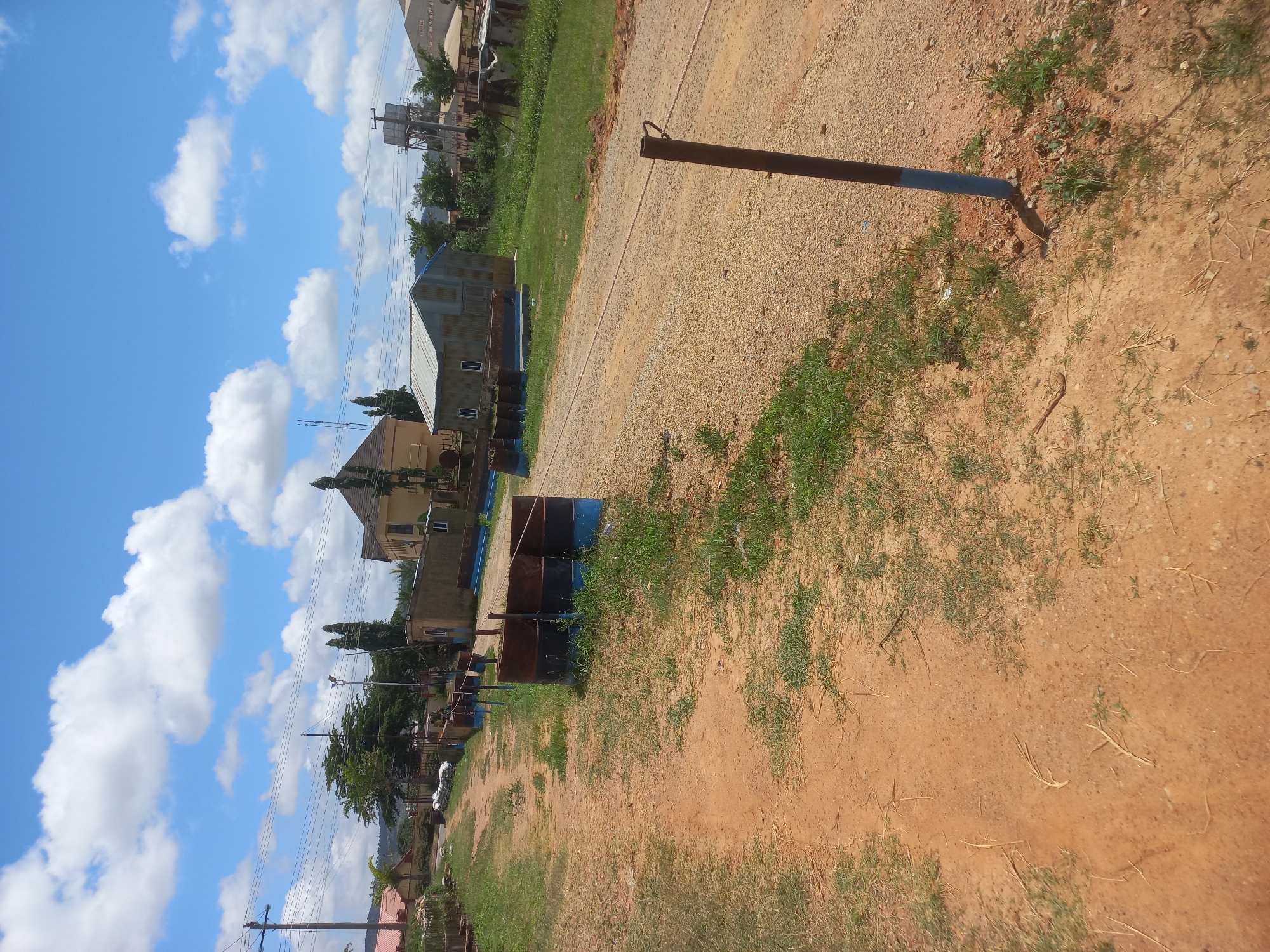 The image depicts an outdoor scene in a rural or developing urban area, likely in New Stadium, Nigeria, under a partly cloudy sky. A dirt path or unpaved road occupies the foreground, with sparse green grass growing along its edges and in patches on the path itself.
Along the left side of the path, several rusty metal barrels, painted with blue bands at the bottom, are arranged in a line. A single, thin metal pole with a wire running through it stands on the right side of the path, also painted with a blue band at its base.
In the middle ground, several structures are visible. To the left, a light yellow building with a brown roof stands. Adjacent to it is a smaller, corrugated metal structure with white-framed windows. Further to the right, a larger yellow building with a brown roof is situated. Behind these structures, the landscape features trees, vegetation, and what appears to be a water tower. The background suggests a moderately populated area with buildings and infrastructure.
No people are visible in the image. The overall impression is one of a somewhat undeveloped or industrial area on a bright, sunny day.