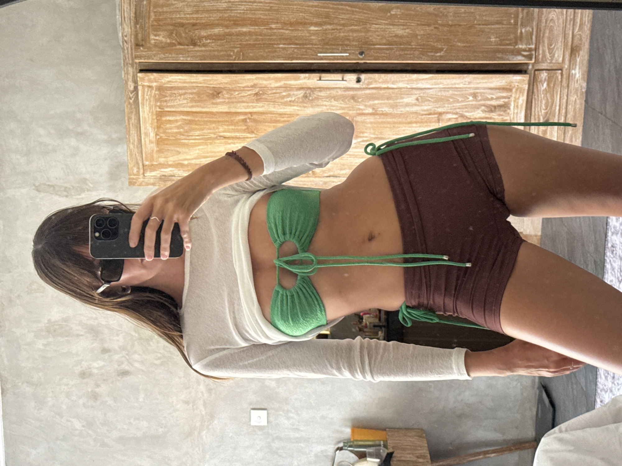 A woman in a green knit bikini top with string ties and brown shorts stands in front of a mirror, taking a selfie with a black smartphone. She wears a long-sleeved white top and sunglasses. A wooden cabinet is behind her, and the scene appears to be indoors, possibly a bathroom or dressing room in North Kuta, Indonesia.