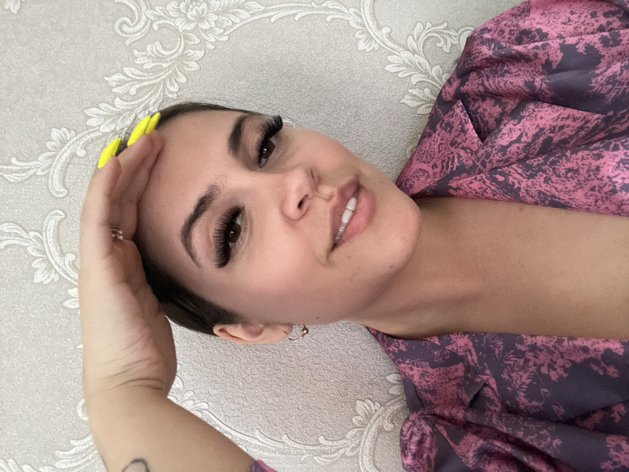 A tilted, close-up indoor portrait features a young woman with dark hair and a light complexion, looking towards the camera with a soft smile. Her head is angled, and she appears to be lying down or reclining. She has pronounced, dark eyelashes and well-defined eyebrows. Her left hand is visible, resting near her forehead, showcasing long, neon yellow fingernails and a ring on her ring finger. A dark, partial tattoo is visible on her left forearm. A small, golden hoop earring adorns her right ear.
She is dressed in a garment that features a vibrant, mottled pattern in shades of dark pink and purple, possibly a robe or patterned top. The background is a light grey wall adorned with a classic white and light grey floral or damask pattern. The image conveys a relaxed and pleasant mood, likely a self-portrait. No text is visible in the image. The location context is Chaikovsky, Russia.
