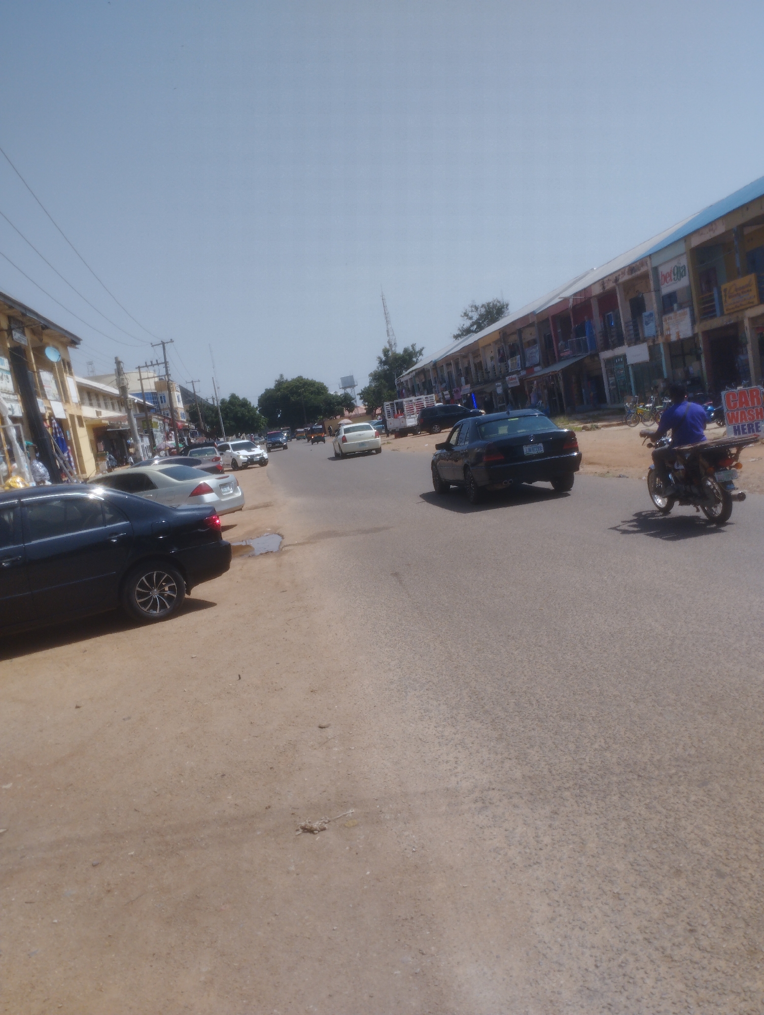 The image captures a daytime scene on a street in Jos, Nigeria. The sky is clear and bright, suggesting fair weather. The street is lined with buildings that appear to be shops or commercial establishments, with awnings and signage indicating various businesses.
In the foreground, a dark sedan is parked on the side of the road, with its rear facing the camera. To the right of the sedan, the street surface is a mix of asphalt and dirt, with a small puddle of water. Further down the street, several other cars are parked or in motion. A white sedan is visible, along with a darker vehicle.
In the middle ground, a black car is driving on the road. To the right, a person on a motorcycle is riding towards the camera. A prominent sign with red and white lettering is visible, reading "CAR WASH HERE".
The background reveals more buildings and trees, with a communication tower visible in the distance on the left. The overall atmosphere appears to be that of a typical day in a bustling commercial area. The presence of parked cars, shops, and traffic suggests ongoing activity and daily life. The lighting indicates it is likely mid-morning or afternoon.