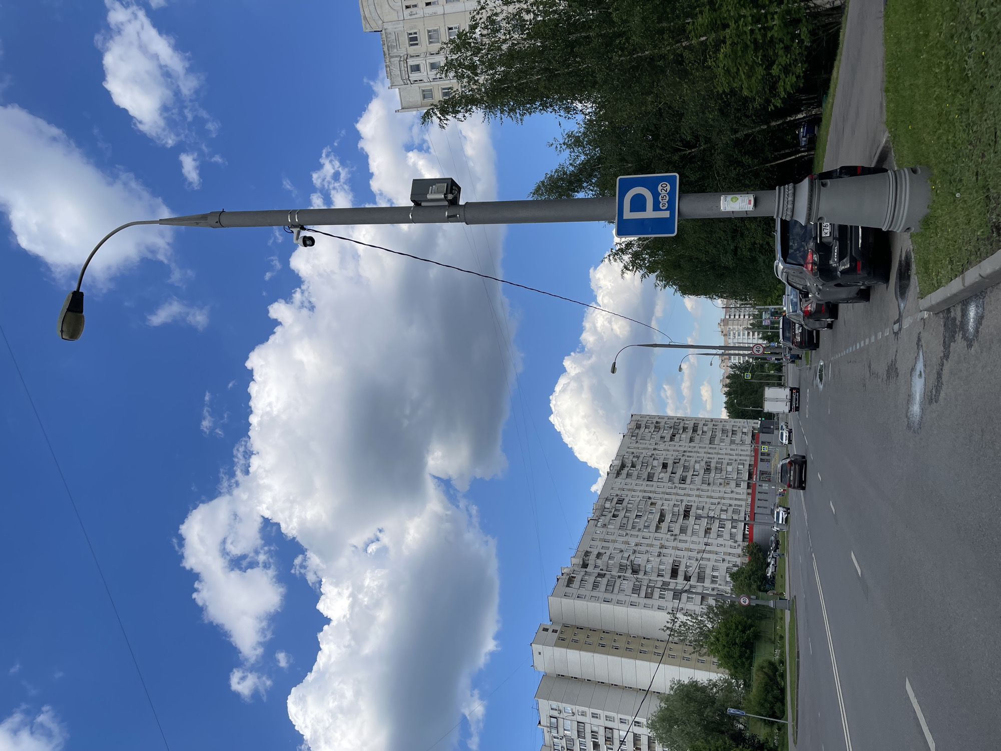 The image captures an urban street scene in Moscow, Russia, under a clear blue sky with scattered white cumulus clouds. The perspective is slightly angled, looking down a straight road.
In the foreground, a tall, grey utility pole stands prominently on the right. A street light fixture curves from the pole towards the left. Attached to the pole are several objects: a blue rectangular parking sign displaying a white "P" symbol with the numbers "10 15 20" beneath it, a black rectangular device (possibly a camera or sensor) mounted high up, and another smaller camera or sensor lower down. Wires extend from the pole. The base of the pole features a decorative, fluted grey cover.
Along the right side of the road, several cars are parked, facing away from the viewer. One visible license plate reads "K76" (partially obscured). The road itself is asphalt, featuring white lane markings and some wet patches or puddles. Further down the road, more cars are visible, both parked and in motion. A speed limit sign indicating "40" (likely 40 km/h) is faintly visible in the distance.
On the left side of the image, multiple large residential buildings with numerous windows and balconies rise into the sky. Some buildings appear older with a beige or light grey facade, while others are taller and more modern in design. Green trees and other vegetation line the roadside, separating the buildings from the street and casting shadows.
The scene depicts a typical bright day, likely midday or afternoon, given the clear illumination. The overall impression is one of a busy urban environment with a mix of residential structures and everyday street activity.