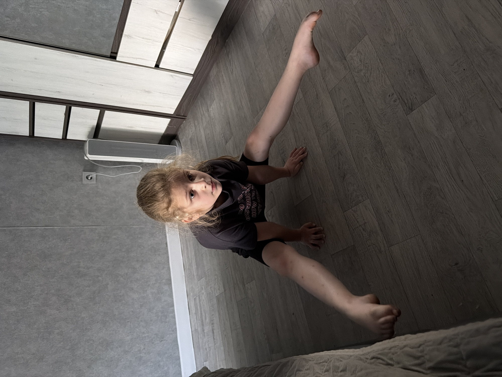 A young girl with blonde hair is sitting on a wooden floor, her body is oriented towards the left of the frame. She is wearing a black t-shirt and black shorts. Her right leg is extended upwards and slightly to the left, while her left leg is bent and tucked towards her body. Her hands are resting on the floor. She is looking at the camera with a neutral expression. The scene is indoors, with a wall and cabinetry visible in the background. The lighting suggests it may be daytime, but the specific time is not determinable. There is a white vent on the wall behind her. The floor is made of grey wood-look laminate planks. The overall mood is casual and candid. The t-shirt has some visible text which appears to be "DO YOU INSTRUCT HOW TO..." but the rest is obscured.