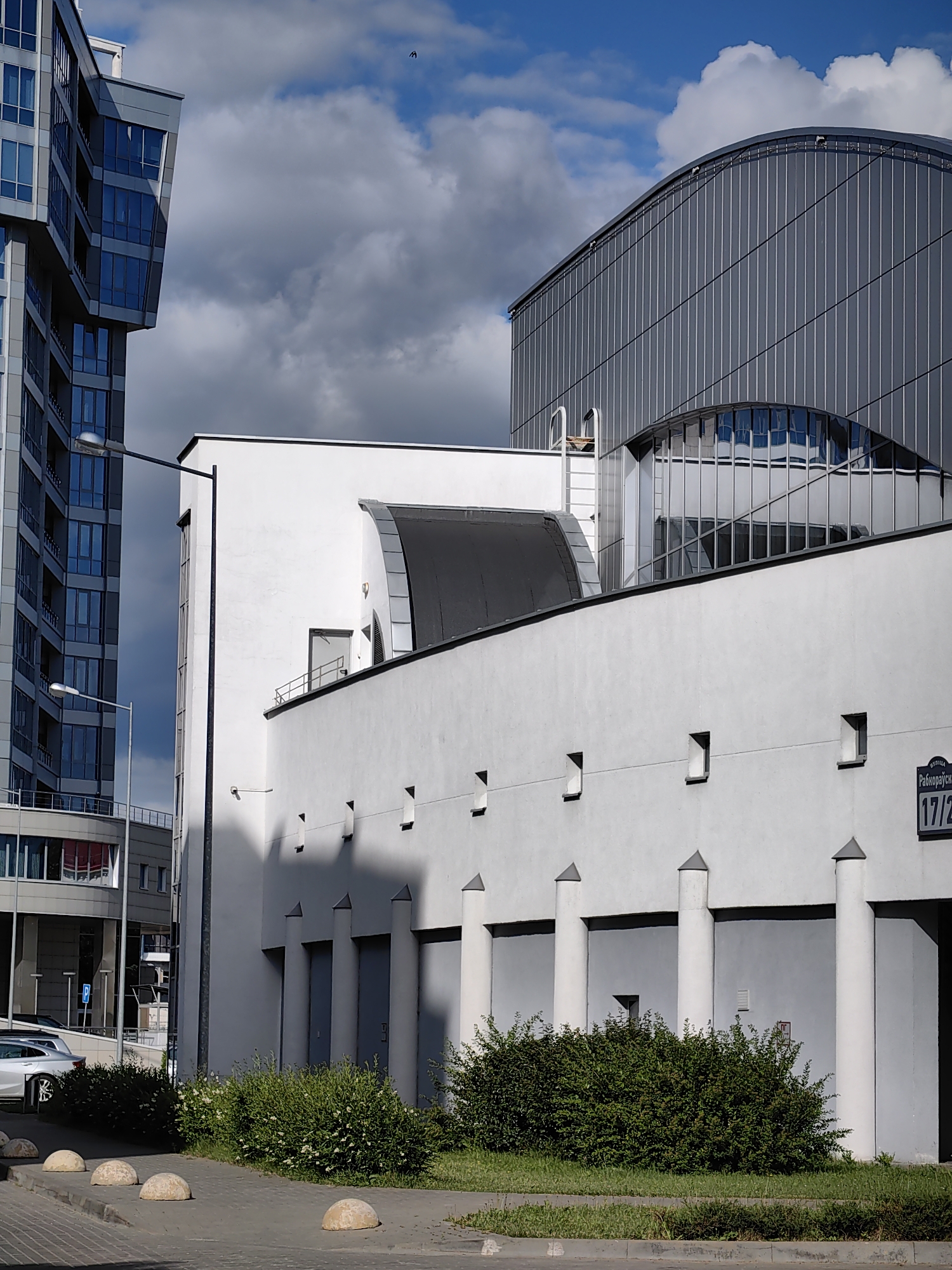The image depicts a street scene in Minsk, Belarus, with a focus on modern architecture and urban elements. On the left, a tall, multi-story building with a geometric design and blue-tinted windows stands prominently. To its right, a white, low-rise building with a rounded, gray-paneled roofline dominates the center of the frame. This building features a series of evenly spaced, dark gray doors and white cylindrical columns extending from its facade. Small, square openings are visible on the upper part of the white wall, casting shadows. A street sign with Cyrillic text and the numbers "17/2" is affixed to the right side of this building. In the foreground, a paved sidewalk is lined with spherical concrete bollards. A patch of green grass and dense shrubbery with small white flowers grows along the edge of the sidewalk. Several cars are parked on the street to the left. The sky above is a mix of bright blue and dramatic dark clouds, suggesting dynamic weather conditions. The scene is captured on a sunny day, with strong shadows indicating the position of the sun.