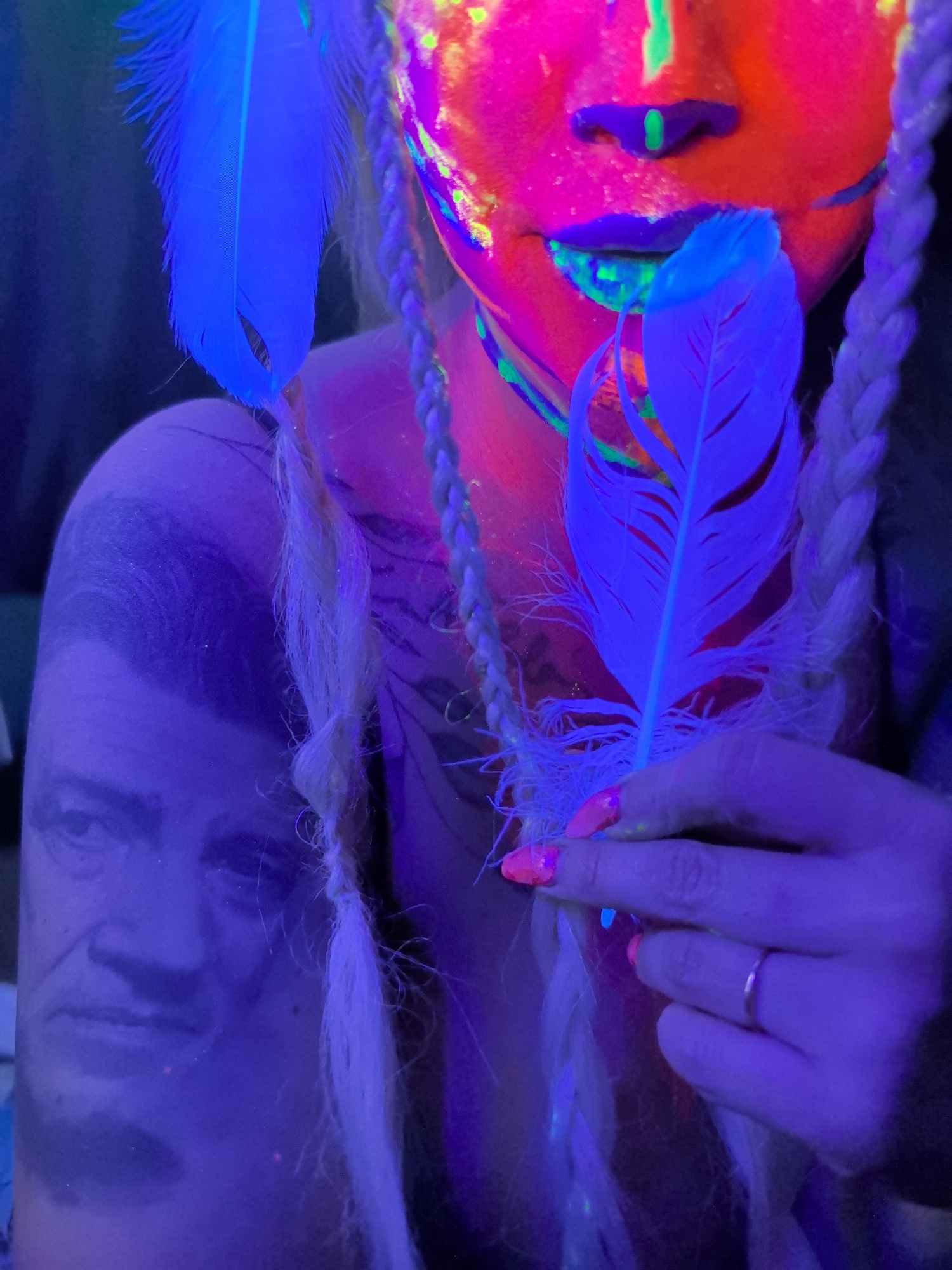 An artistic portrait taken under blacklight shows a person adorned with fluorescent paint and glowing objects. The subject's face and upper body are painted with vibrant neon pink, green, and blue colors, which glow intensely under the UV light. Two prominent, glowing blue feathers are featured, one held by the person's hand near their mouth, and another positioned to the left side of the frame.
The person has long, light-colored hair styled into braids, which also appear to glow. On the left arm, a large portrait tattoo of an older man is visible, appearing in hues of purple and blue due to the UV lighting. The person's hand is visible, featuring bright, fluorescent pink nail polish and a simple metallic ring on one finger. The background is dark and indistinct, emphasizing the glowing elements. The overall scene creates a vibrant, artistic, and otherworldly atmosphere. No discernible text is visible in the image.