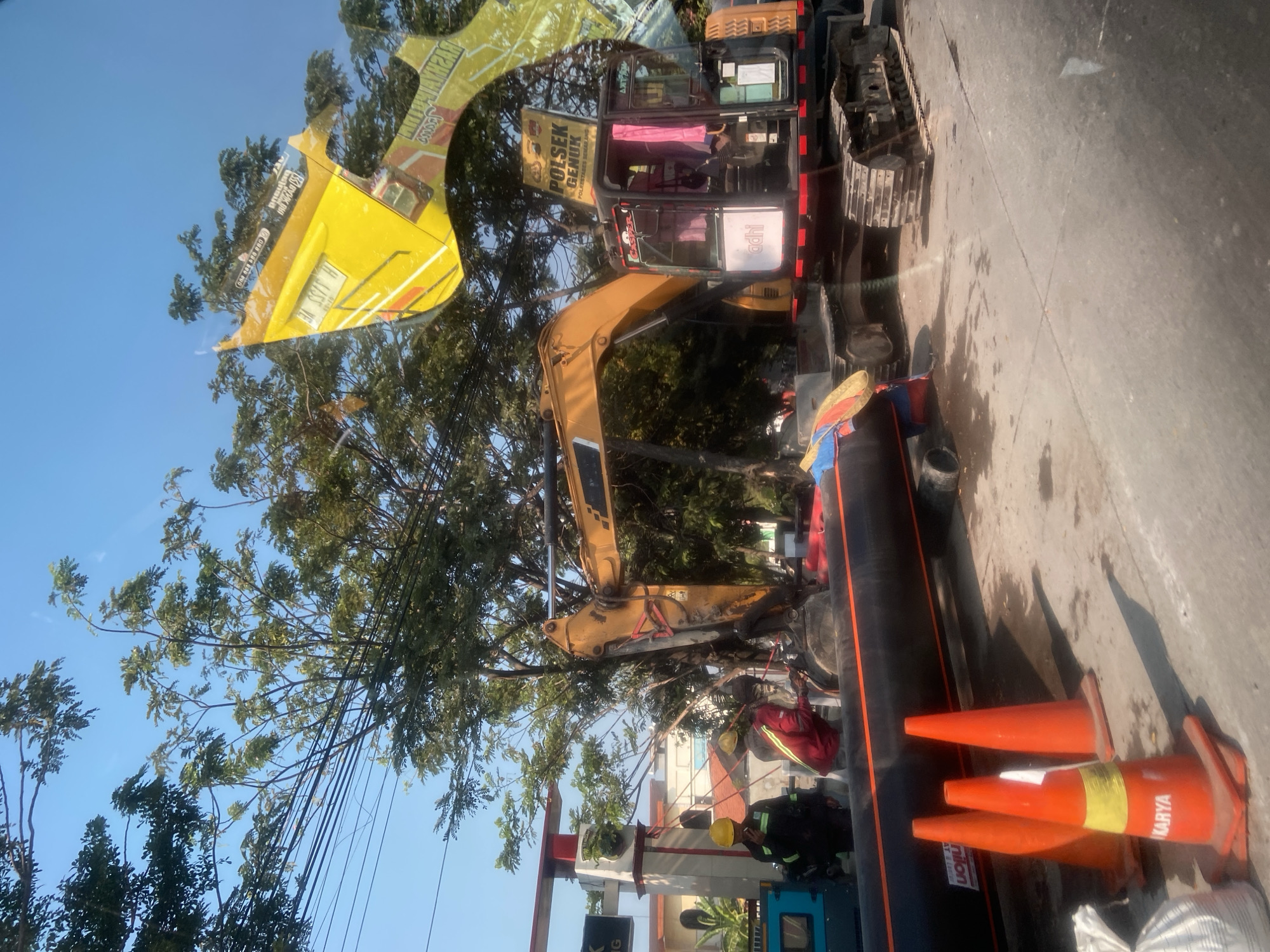 An excavator is positioned on a paved road in Semarang, Indonesia, in front of a building with a red and white sign. Two construction workers, one in a yellow hard hat and dark uniform, the other in a red jacket and yellow hard hat, are near the excavator, which has large black pipes and orange traffic cones in the foreground. The excavator's cabin is visible, with pink curtains inside. A yellow banner with text is draped over the excavator's boom. A blue container is to the left of the excavator. The sky is clear and blue, with trees and power lines overhead.