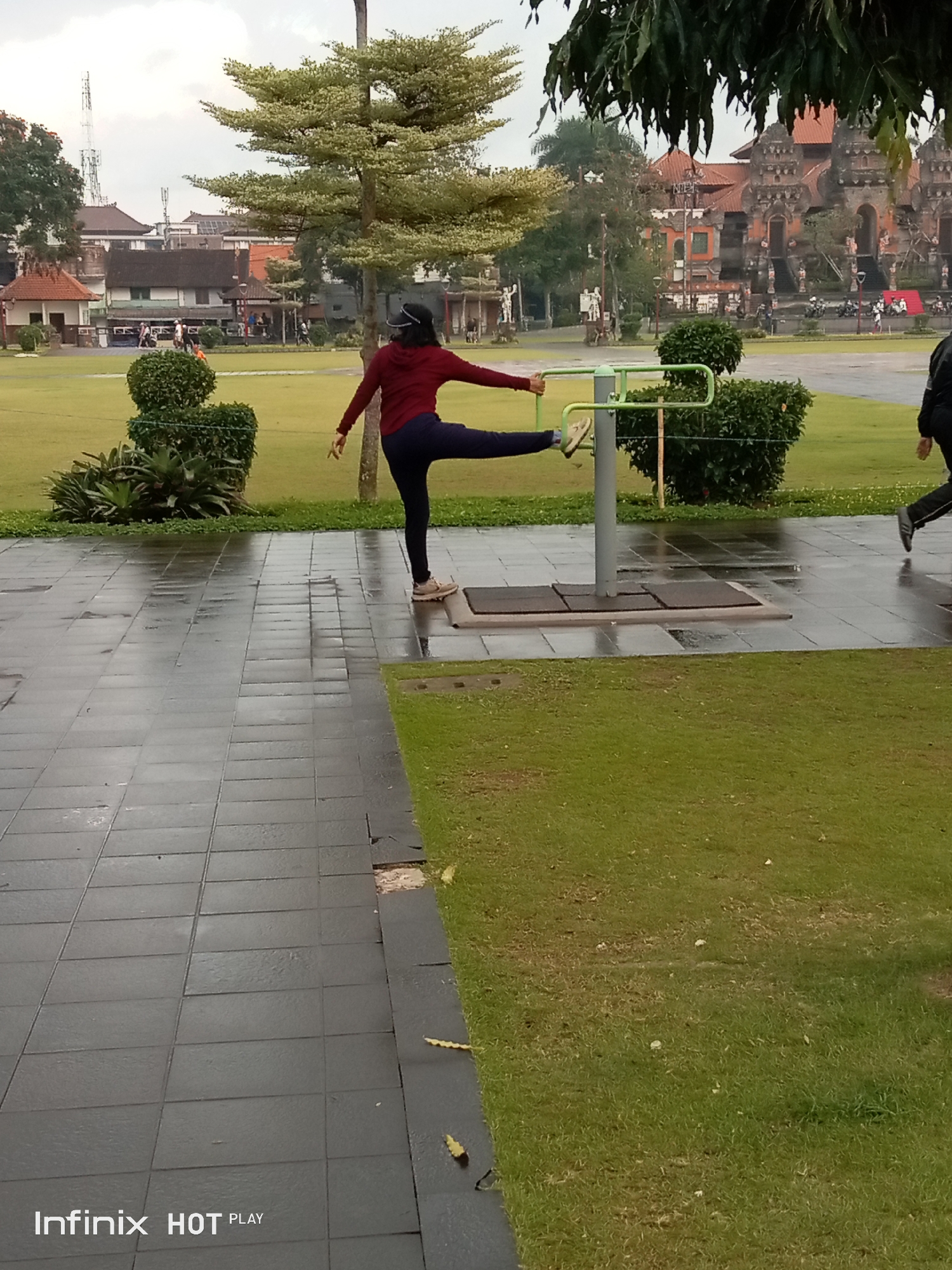 The image depicts an outdoor scene, likely a park or public recreational area, in Gianyar, Indonesia. In the foreground, a person wearing a dark red top and dark blue pants is engaged in a leg exercise on a piece of outdoor fitness equipment. The equipment consists of a central pole with horizontal bars, and the person is holding onto one of the bars with their left hand while extending their right leg forward and upwards. Their right foot is resting on the ground for balance. The ground in the foreground is paved with dark gray square tiles that appear wet, suggesting recent rain or cleaning. Adjacent to the paved area is a well-maintained green lawn.
In the middle ground, more green grass is visible, along with decorative shrubs. Further in the background, a traditional Balinese temple-like structure with intricate stone carvings and orange tiled roofs is prominent. Several trees are scattered throughout the scene, some with light-colored foliage. In the distance, buildings and a radio tower can be seen. Another person, wearing dark clothing, is captured in motion, running towards the right side of the frame. The sky is overcast, indicating a cloudy day. The overall impression is of a public space being utilized for recreation and featuring elements of local architecture.