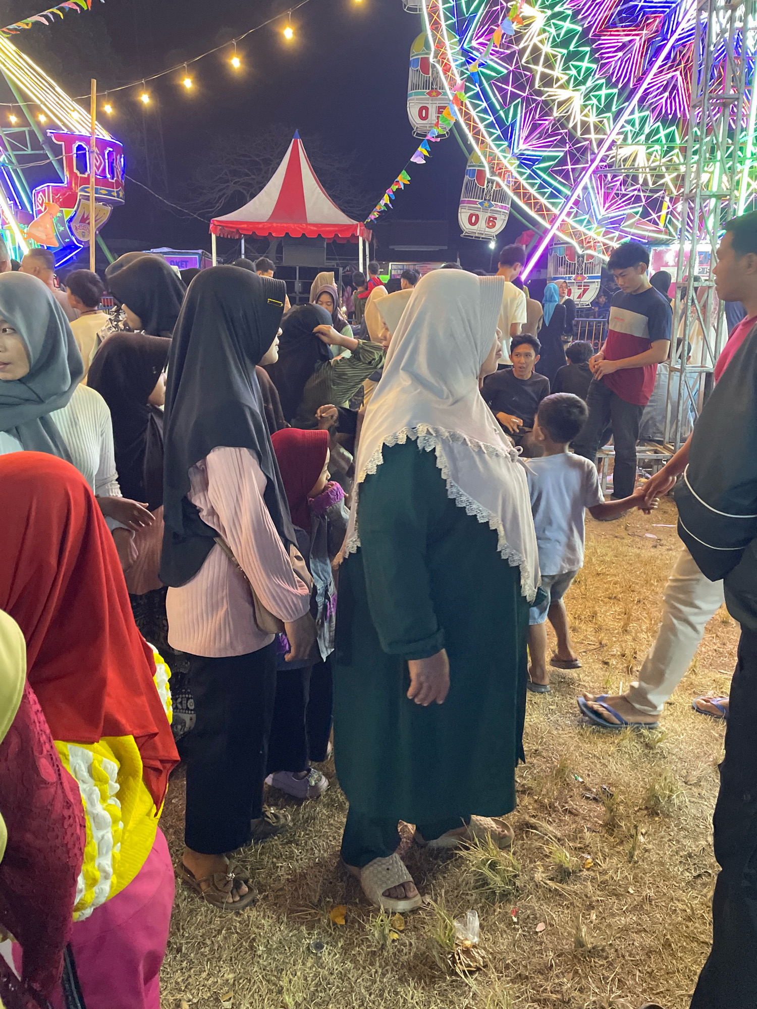 The image depicts a nighttime scene at what appears to be a local fair or carnival in Pulung Kencana, Indonesia. A Ferris wheel with vibrant, multicolored lights dominates the background, illuminating the surrounding area. In the foreground and midground, a crowd of people, predominantly women and children dressed in traditional Islamic clothing including hijabs and long dresses, are gathered. Some individuals are holding hands, suggesting familial or friendly connections. The ground is covered with dry grass and scattered debris. Other elements visible include a striped tent, string lights, and potentially food stalls or game booths, typical of a carnival setting. The overall atmosphere suggests a lively community gathering.