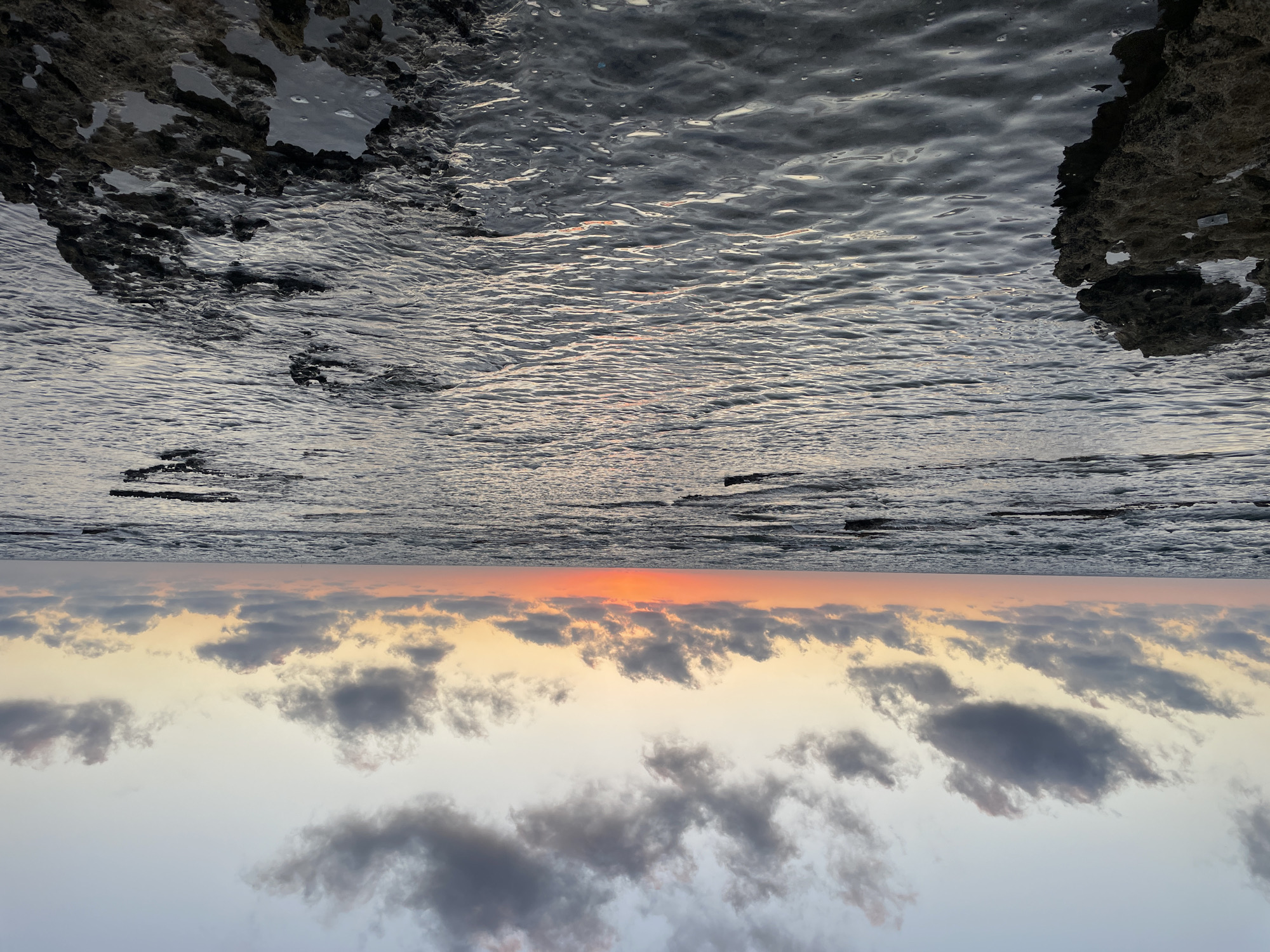 The image captures a sunset over the Mediterranean Sea, as seen from the rocky coast of Haifa, Israel. The scene is divided horizontally by the horizon. The upper portion of the image depicts the sea and the rocky shore. The water is slightly choppy, reflecting the dimming light of the sky. Dark, textured rocks jut out from the left and right sides, framing the view of the water. In the lower portion, the sky transitions from a warm, soft orange glow near the horizon, indicative of the setting sun, to a pale, grayish-blue at the top. Scattered, dark clouds drift across the sky, their undersides catching the last rays of sunlight. The overall mood is serene and peaceful, with the natural beauty of the coastal landscape at dusk. There is no visible text in the image.