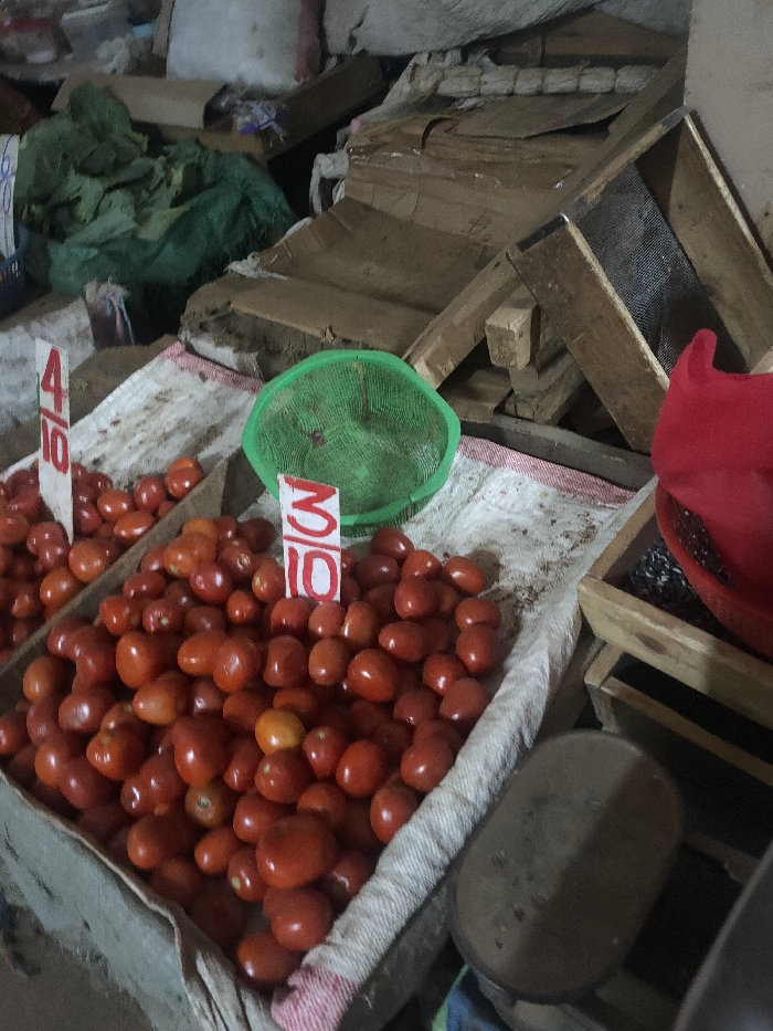 This image depicts a market stall in Ngong ward, Kenya, focusing on produce display. The foreground features two wooden crates filled with numerous ripe, red tomatoes. Both crates are lined with a light-colored, possibly off-white, fabric that exhibits red stripes at the bottom edge. Price markers are visible: one sign in the left crate reads "4/10" in red text on a white background, indicating a quantity-to-price ratio. A similar sign in the right crate reads "3/10." A green, circular plastic sieve or shallow basket is positioned between the two tomato-filled crates. To the immediate right, a portion of a red fabric item, possibly a bag or covering, is visible within a wooden frame or another crate. The background is composed of a cluttered arrangement of stacked and disarranged wooden planks, disassembled wooden crates, and brown cardboard boxes. A large green sack or tarp containing an undefined bulky item is also present in the upper left background. No individuals, actions, or interactions are visible in the frame. The overall lighting is dim, indicating an indoor or covered market environment.