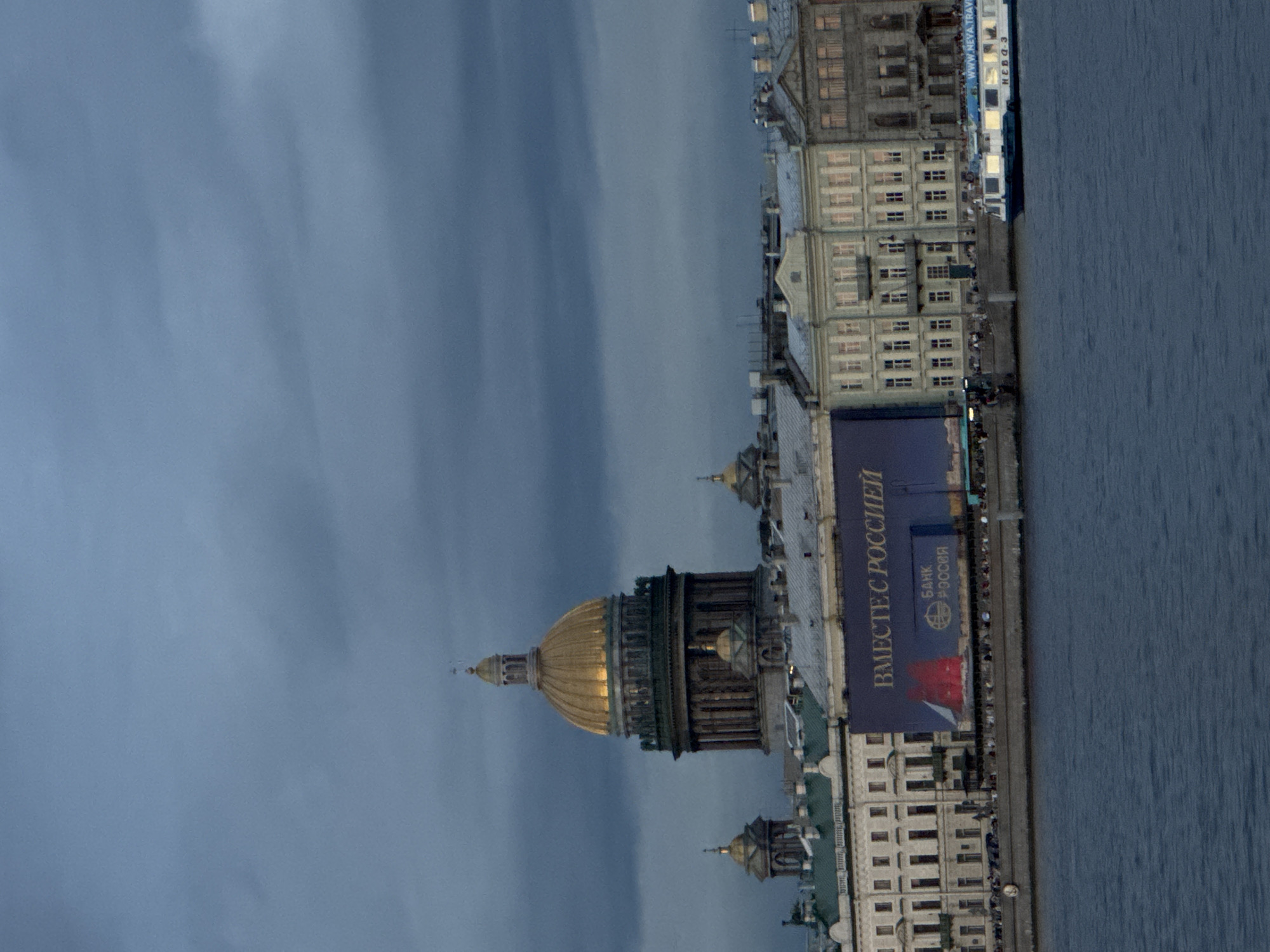 The image captures a view of the Neva River in Saint Petersburg, Russia, during what appears to be twilight or overcast conditions. The most prominent structure is the gilded dome of Saint Isaac's Cathedral, illuminated from within, dominating the skyline. To the right of the cathedral, a row of classical European-style buildings lines the embankment, some with lit windows, suggesting a bustling urban environment.
In the foreground, the dark blue water of the Neva River reflects the muted colors of the sky and the structures. A white and blue tour boat, labeled "WWW.NEVA.TRAVEL" and "NEVA-3", sails eastward along the river. On the embankment in front of the buildings, numerous people are visible, standing and walking along the waterfront. A large blue banner is draped across the facade of one of the buildings, displaying Cyrillic text and a graphic that appears to be a red sailing ship. The sky is filled with heavy, dark clouds, creating a dramatic atmosphere.