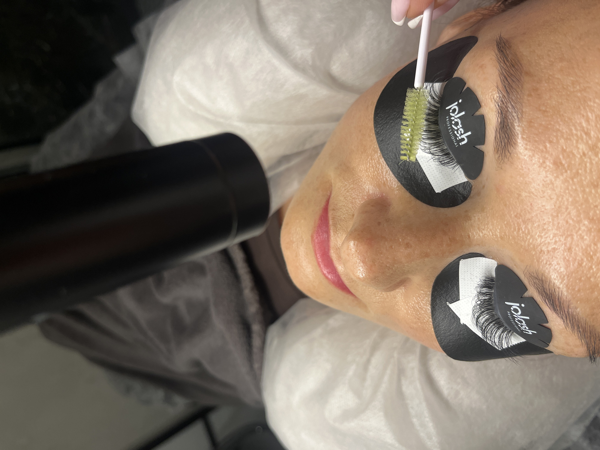 The image shows a close-up, overhead view of a person lying down, undergoing an eyelash extension procedure. The subject's face is tilted upwards, with their eyes closed. Black under-eye patches are applied to both eyes, with one patch on the right eye featuring a black logo that reads "iLash Professional". A green-tipped lash brush is held by a hand with light pink fingernails, and it is positioned near the right eye, appearing to be used to separate or groom the lashes. The person's lips are painted a bright pink color. In the background, there appears to be a dark cylindrical object, possibly a piece of equipment, positioned above the subject's head. The surrounding environment includes white fabric, possibly medical drapes or sheets. There is no visual information in the image that allows for the identification of the location in Białystok, Poland.