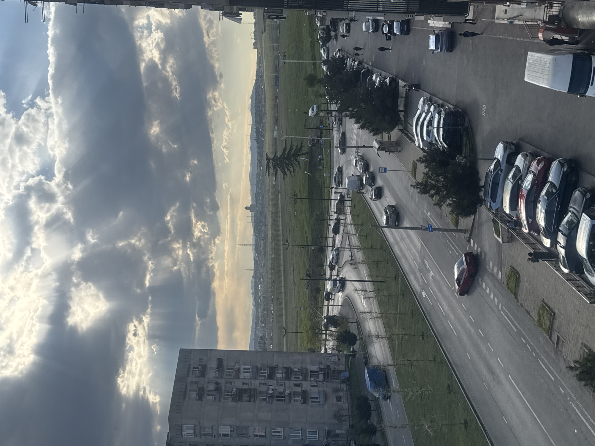 A high-angle view depicts a street scene in Tbilisi, Georgia, characterized by a multilane road with vehicle traffic and parallel parking along the right side. Clouds dominate the sky, with visible sun rays. Adjacent to the road is a large, open green space. Buildings are visible, including a residential building in the lower left corner. Cars are parked and moving along the road. Trees line the street.