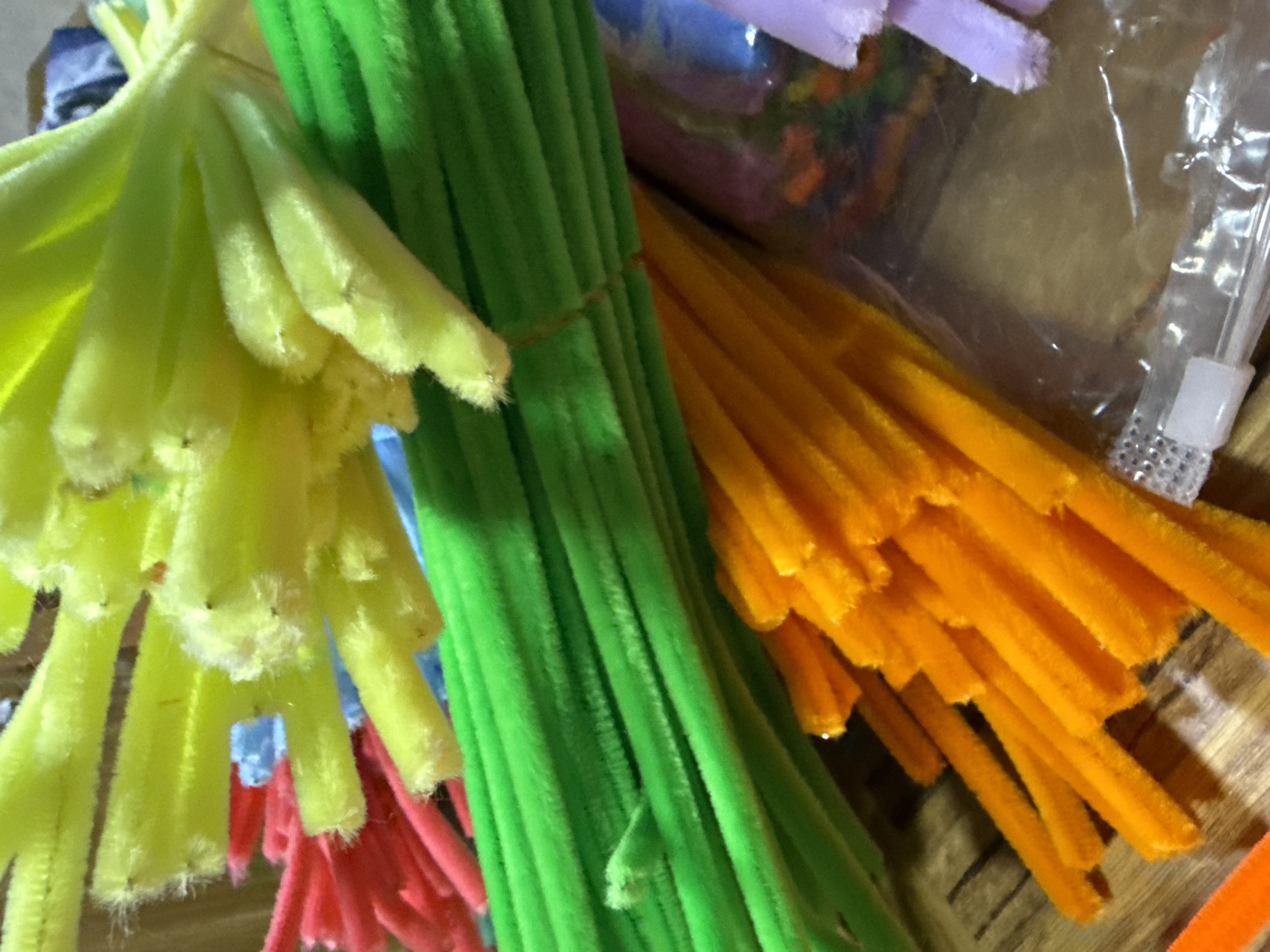 The image presents a close-up view of several bundles of pipe cleaners (chenille stems) in various colors. Dominant colors include yellow, green, orange, and red. The bundles appear to be tightly packed together. A clear plastic bag containing more pipe cleaners of assorted colors is partially visible in the background. A plastic container is visible at the right of the image. The environment appears to be indoors on a wooden or laminate surface. There are no visible indications of the location being in Troyan, Bulgaria.